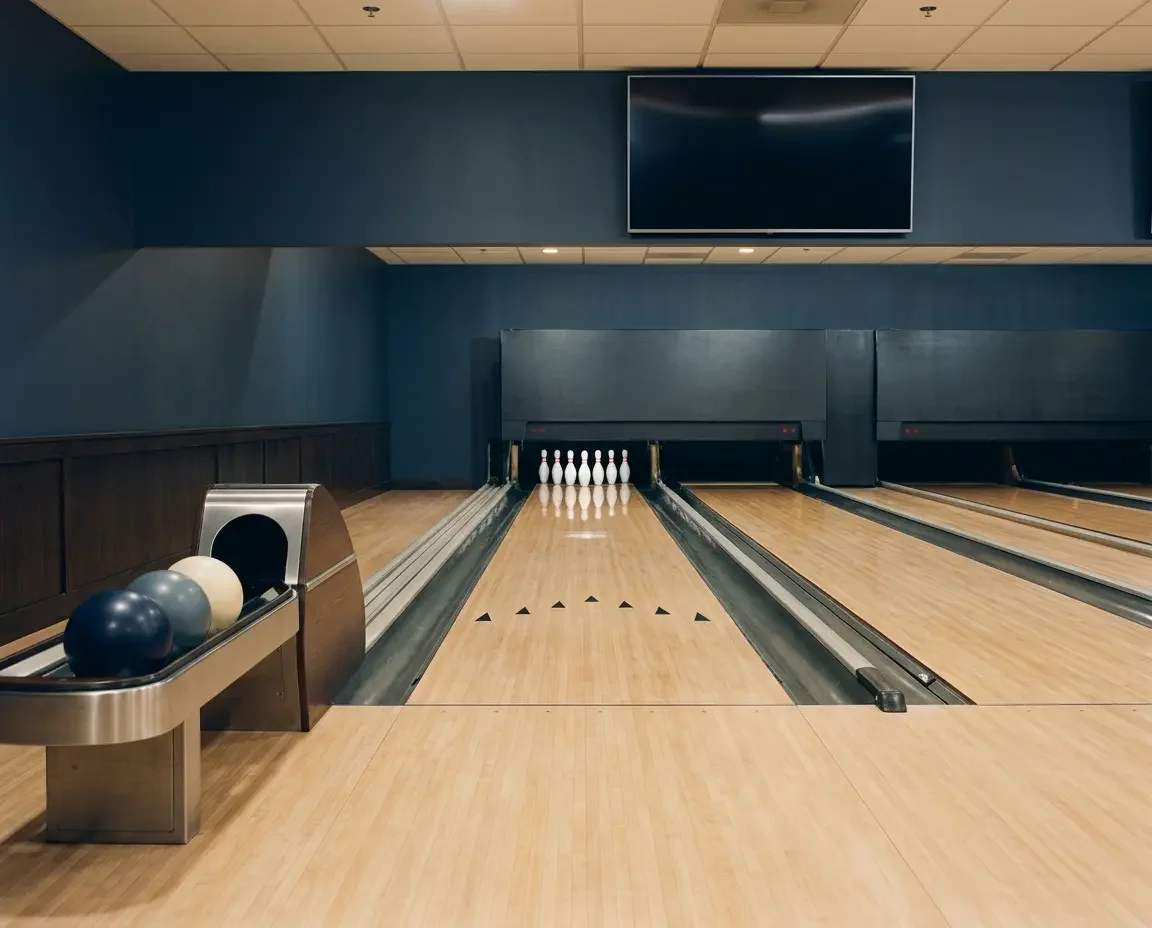 Empty bowling lane with polished maple approach, black targeting arrows, and a full rack of white pins, with three navy, gray, and cream bowling balls in a stainless ball return and a blank wall-mounted screen above a navy back wall.