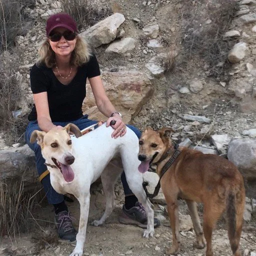 A woman with sunglasses, a hat, and a black shirt outdoors on rocky terrain, sitting and holding two dogs, one white and the other brown.