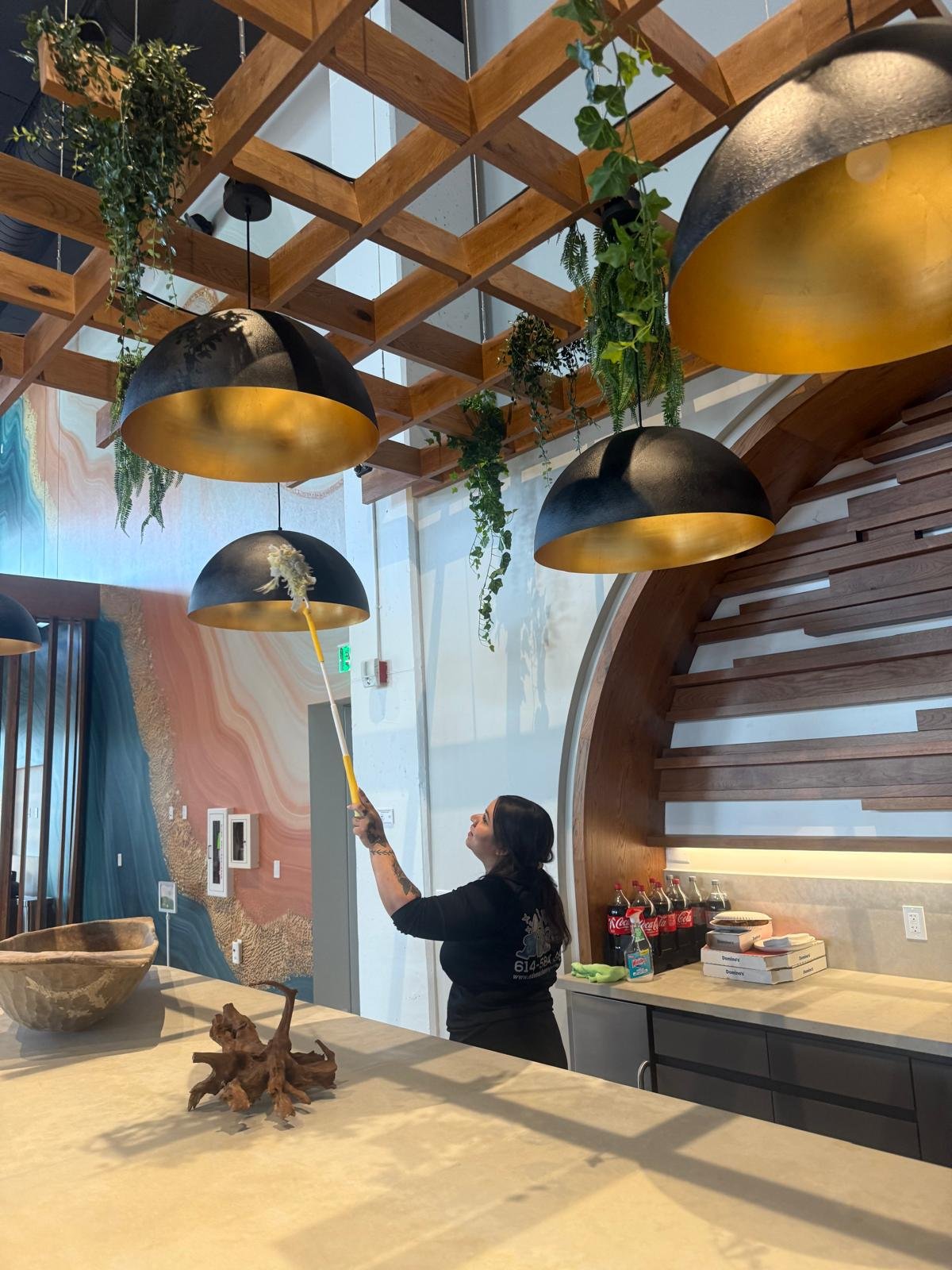 A woman cleaning a black and gold hanging lamp with a duster. The interior has a modern design with green hanging plants, a wooden ceiling structure, and a colorful marble wall. There are soda bottles, pizza boxes, and plates on the counter.