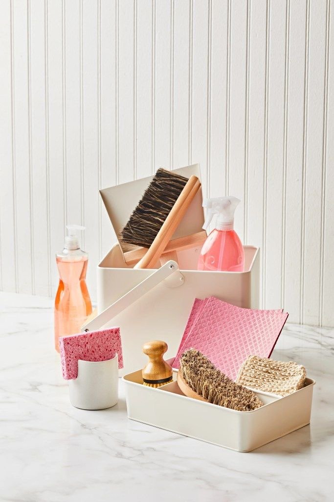 Cleaning supplies including spray bottles, sponges, brushes, and scrubbers organized in white storage containers on a marble countertop with white panel wall background.