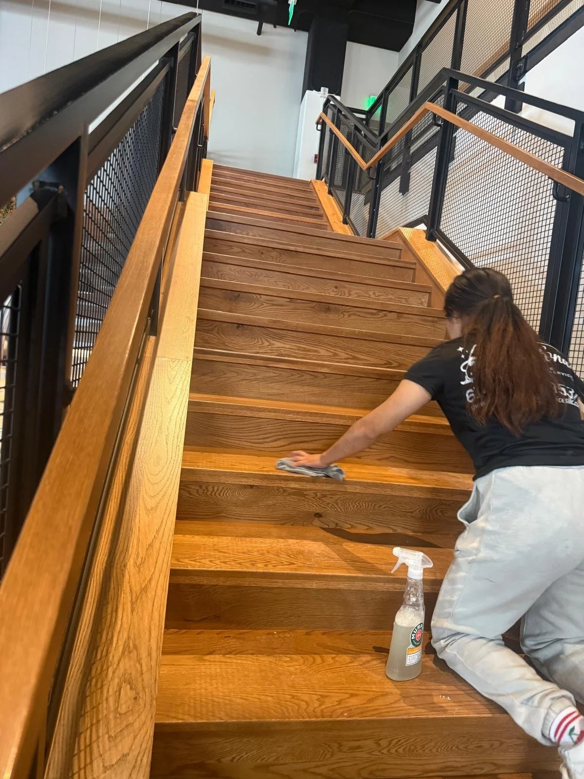 A person cleaning a wooden staircase with a cloth, wearing light-colored pants and a black t-shirt, with cleaning spray bottle on the floor next to them.