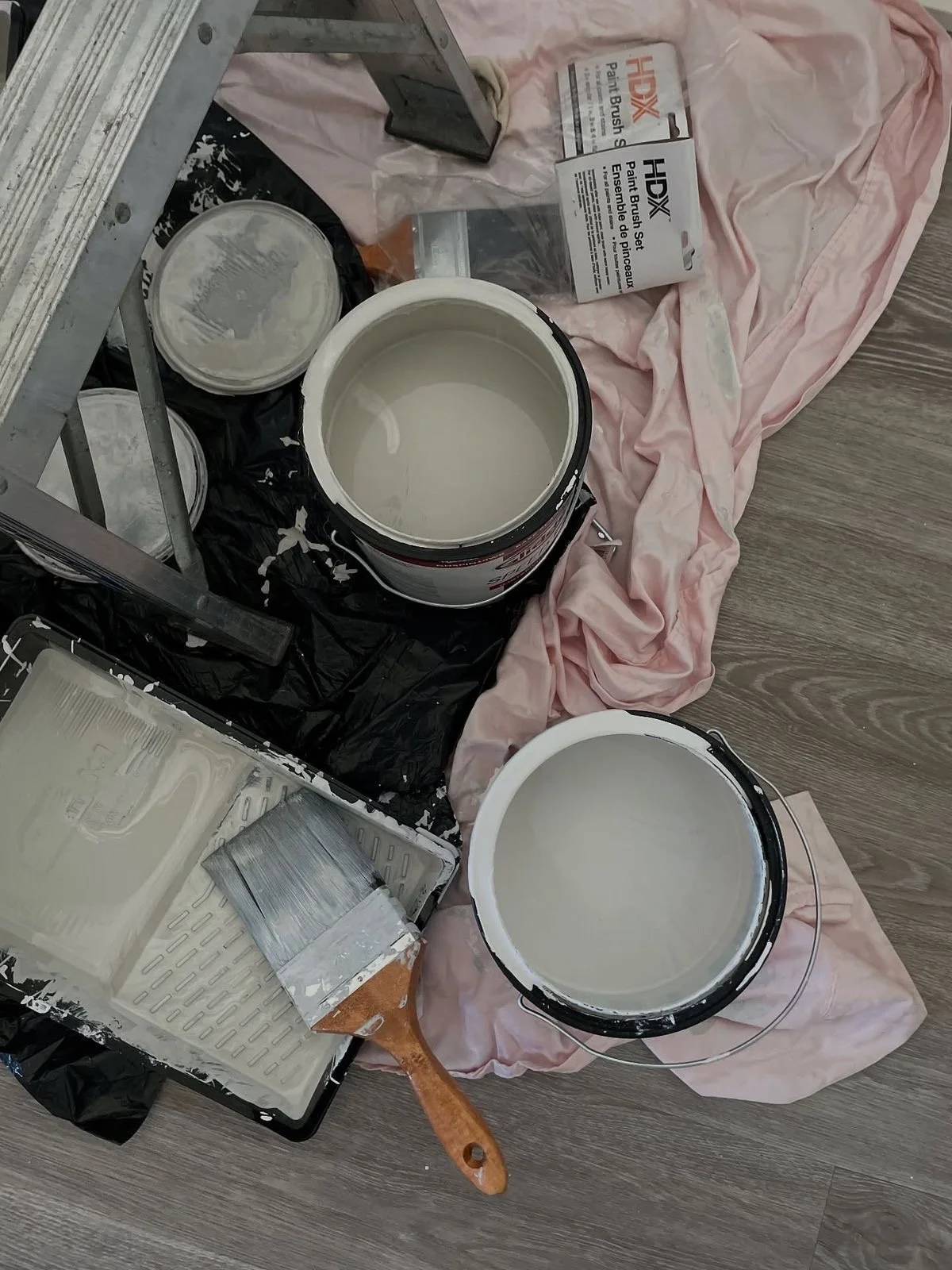 Various painting supplies including two open paint cans with white paint, paintbrush sets in packaging, a small paint roller, and a paint tray, all arranged on a pink cloth and black plastic sheet on a wooden floor.