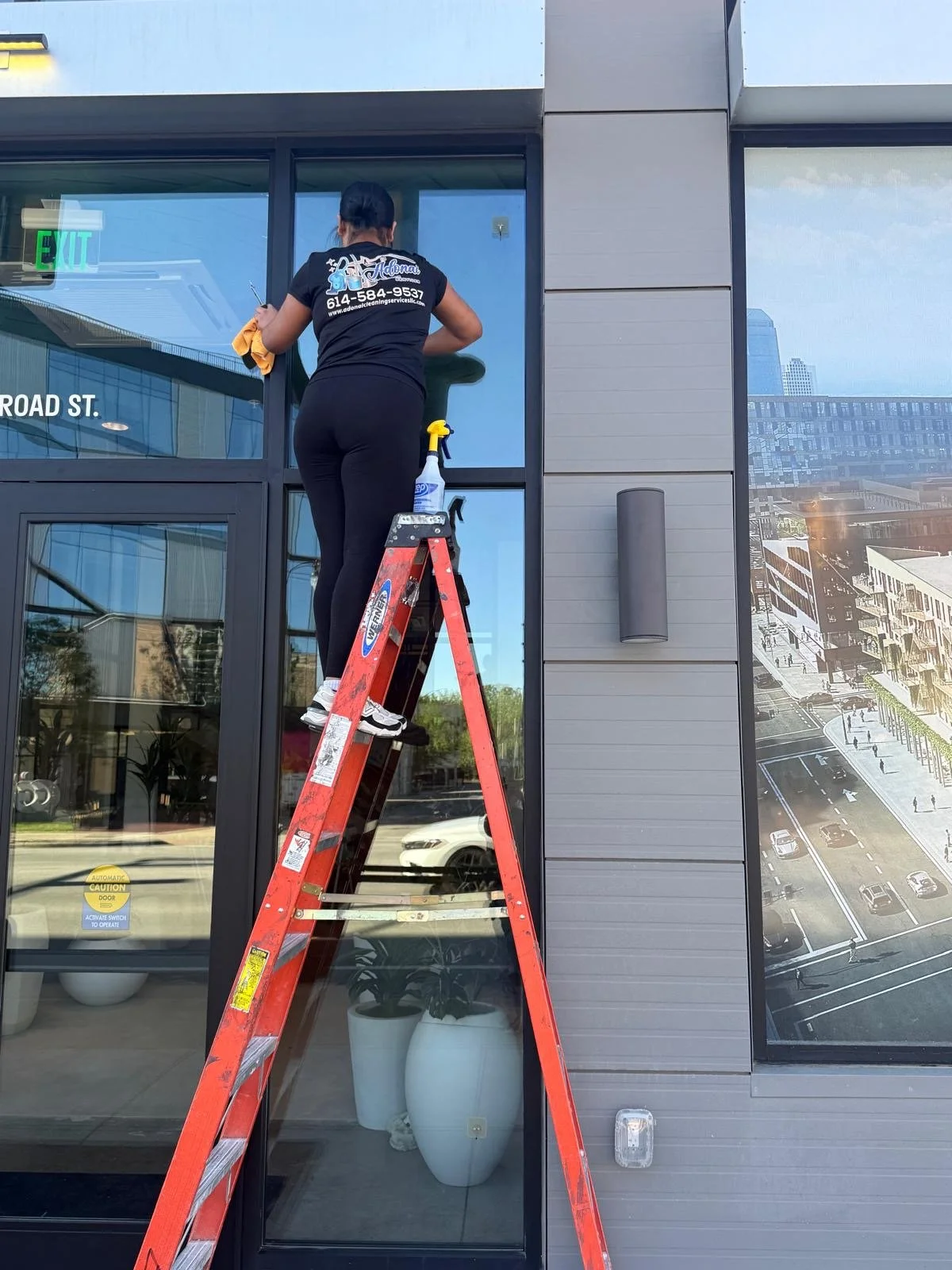 A person cleaning the glass door of a modern commercial building using a yellow cloth, standing on a red step ladder, with cleaning supplies nearby, including a spray bottle. The building features large glass windows and a cityscape reflection, with 