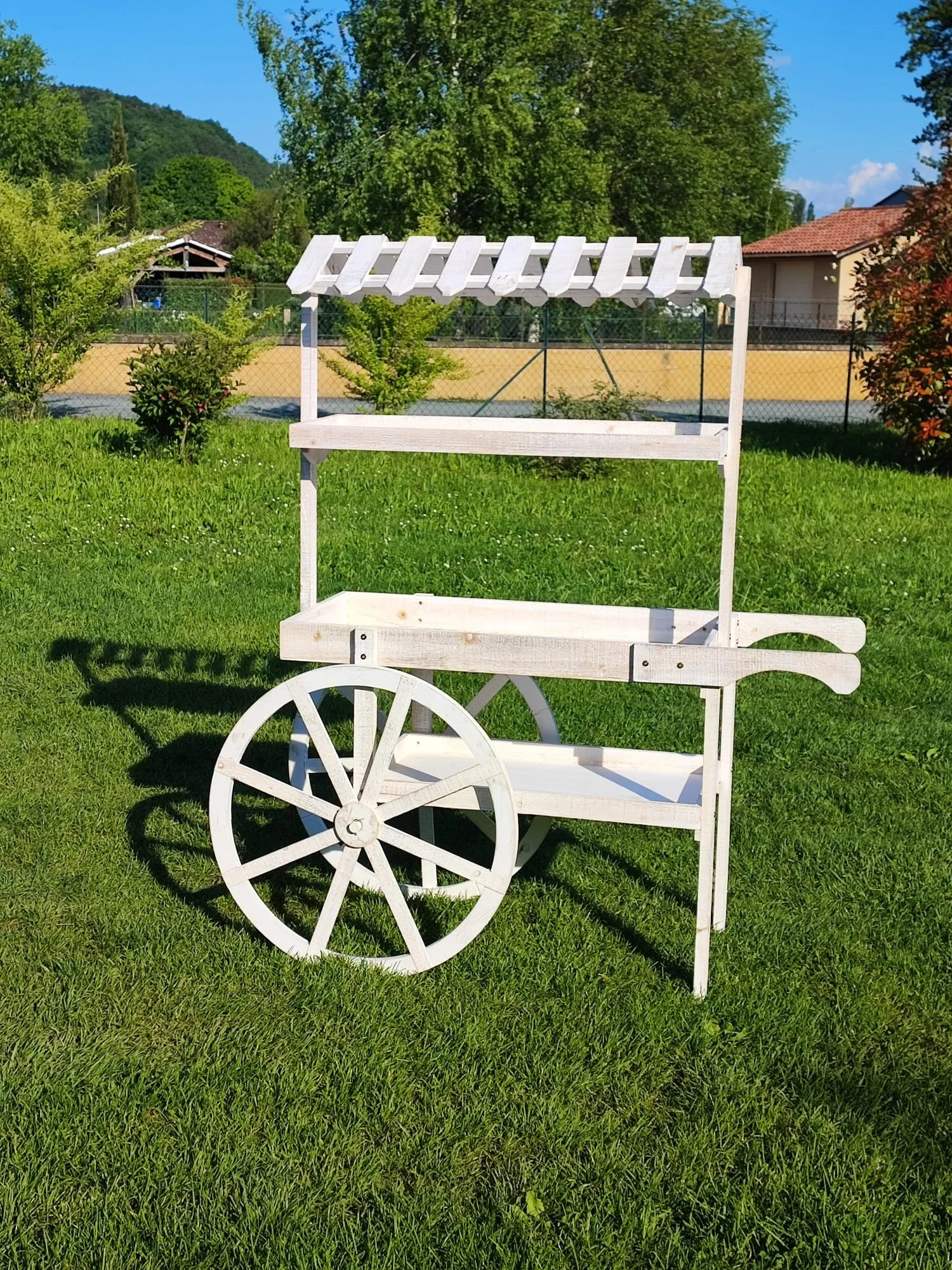 Une carriole en bois blanche dans un jardin verdoyant avec des arbres et des maisons en arrière-plan.