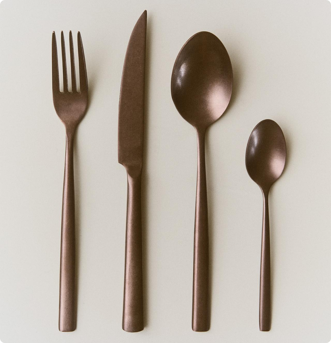 Couverts en métal brossé comprenant une fourchette, un couteau, une grande cuillère et une petite cuillère, tous alignés verticalement sur un fond clair.