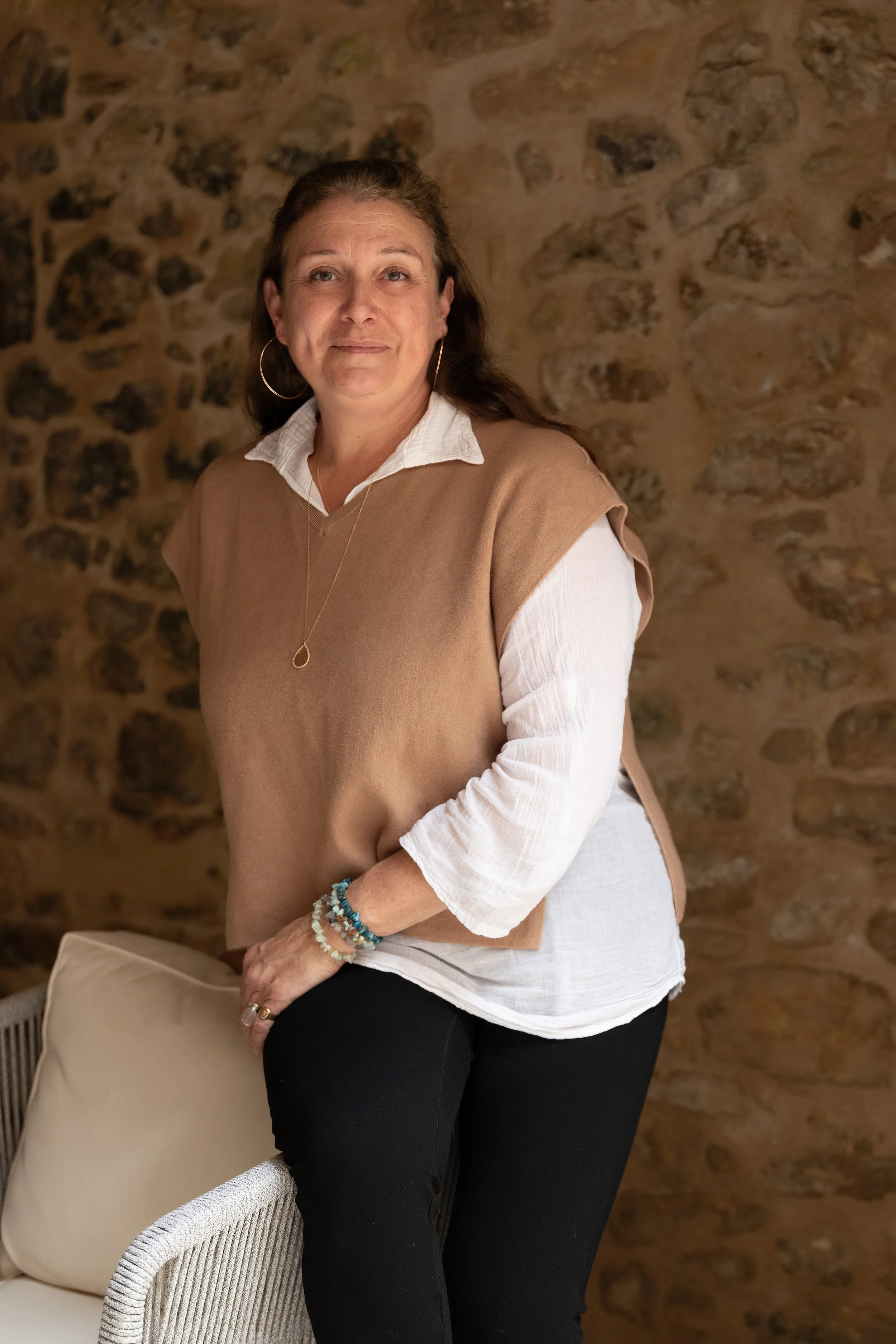 Une femme portant un pull beige, une chemise blanche et des bracelets turquoise, assise à côté d'une chaise en osier, avec un mur en pierre en arrière-plan.