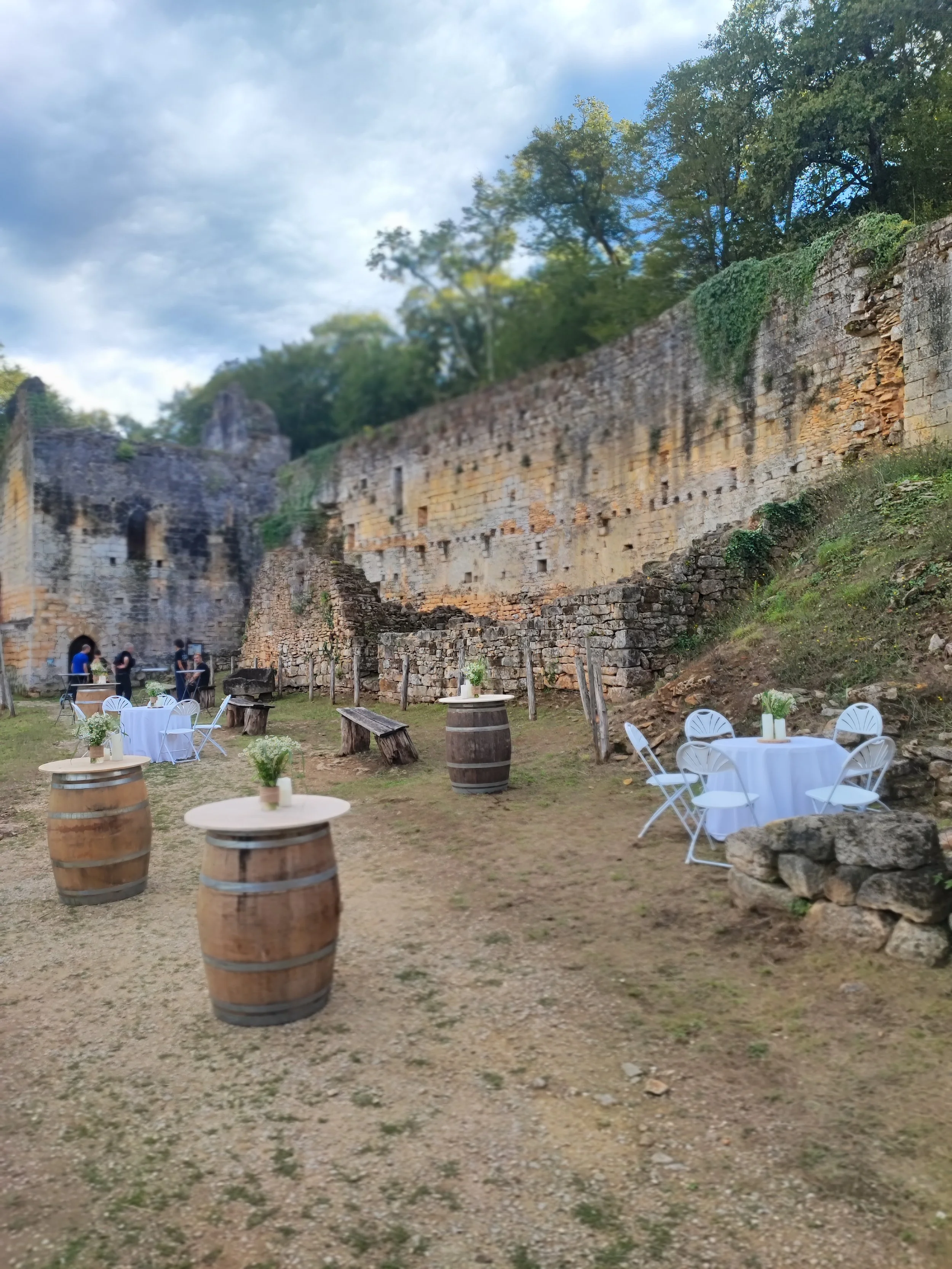 Terrasse en plein air avec des tables rondes couvertes de nappes blanches, des chaises en plastique blanc, des barils en bois utilisés comme tables, un mur en pierre ancien avec des fenêtres et des végétaux en arrière-plan, sous un ciel nuageux.