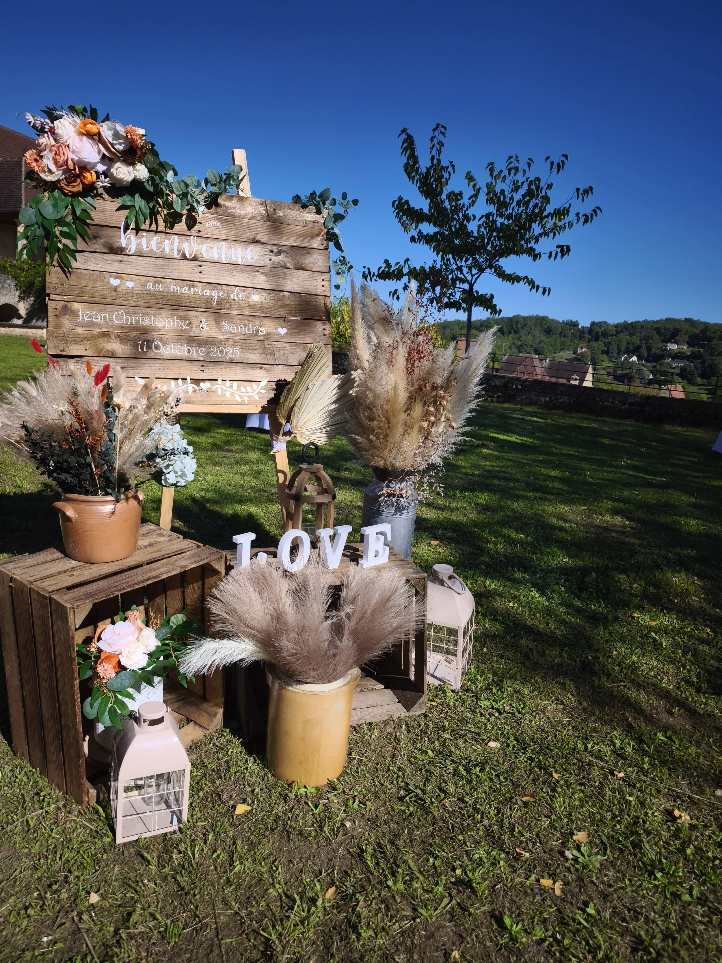 Décoration de mariage en plein air avec un panneau en bois, des fleurs, des lanternes, et des lettres "LOVE" sur l'herbe, sous un ciel bleu.