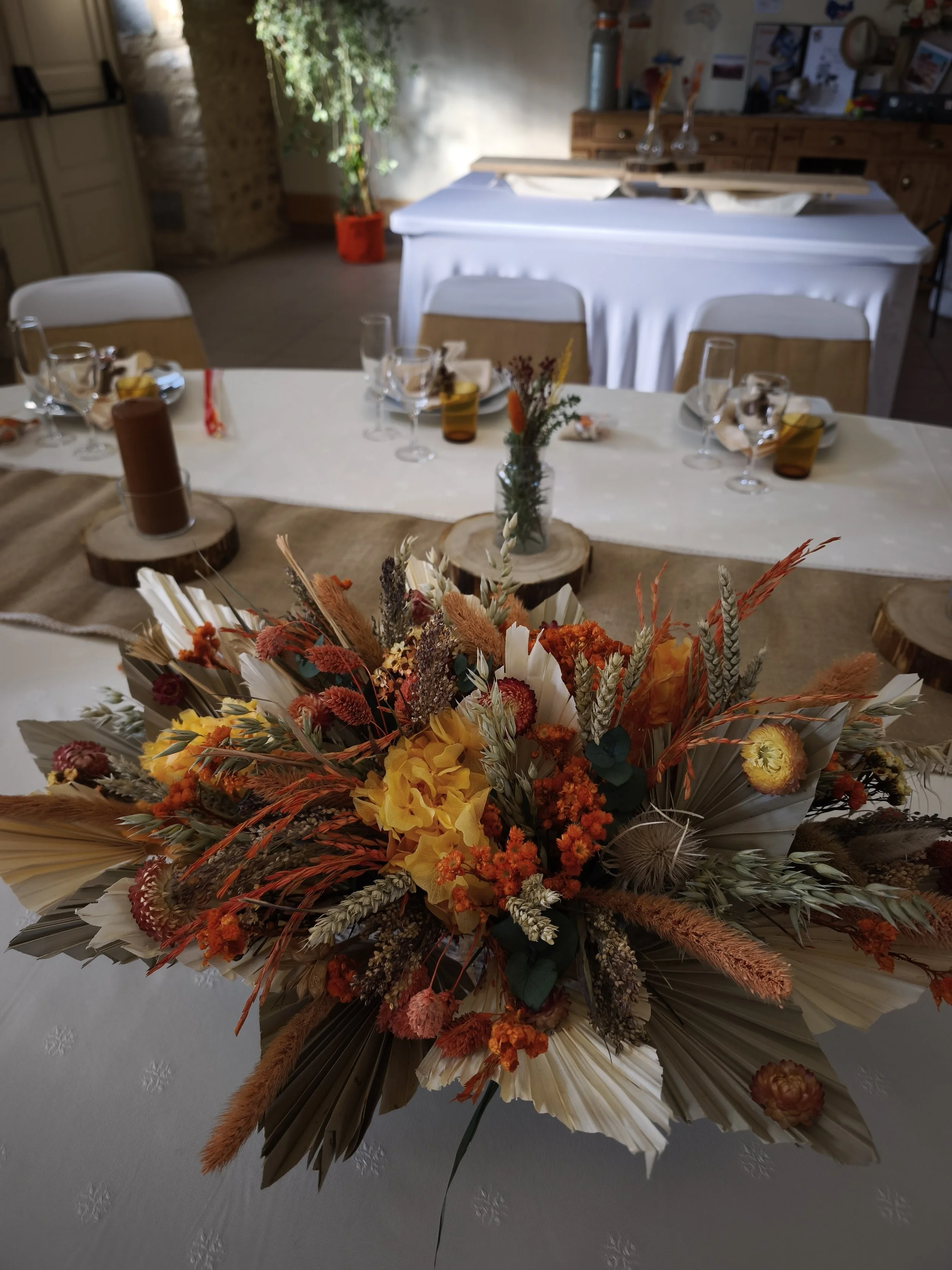 Un centre de table floral composé de fleurs séchées de couleurs jaune, orange, rouge et beige, avec des éléments naturels en papier et en bois, placé sur une table blanche décorée d'une nappe beige. En arrière-plan, une pièce avec un meuble en bois, 