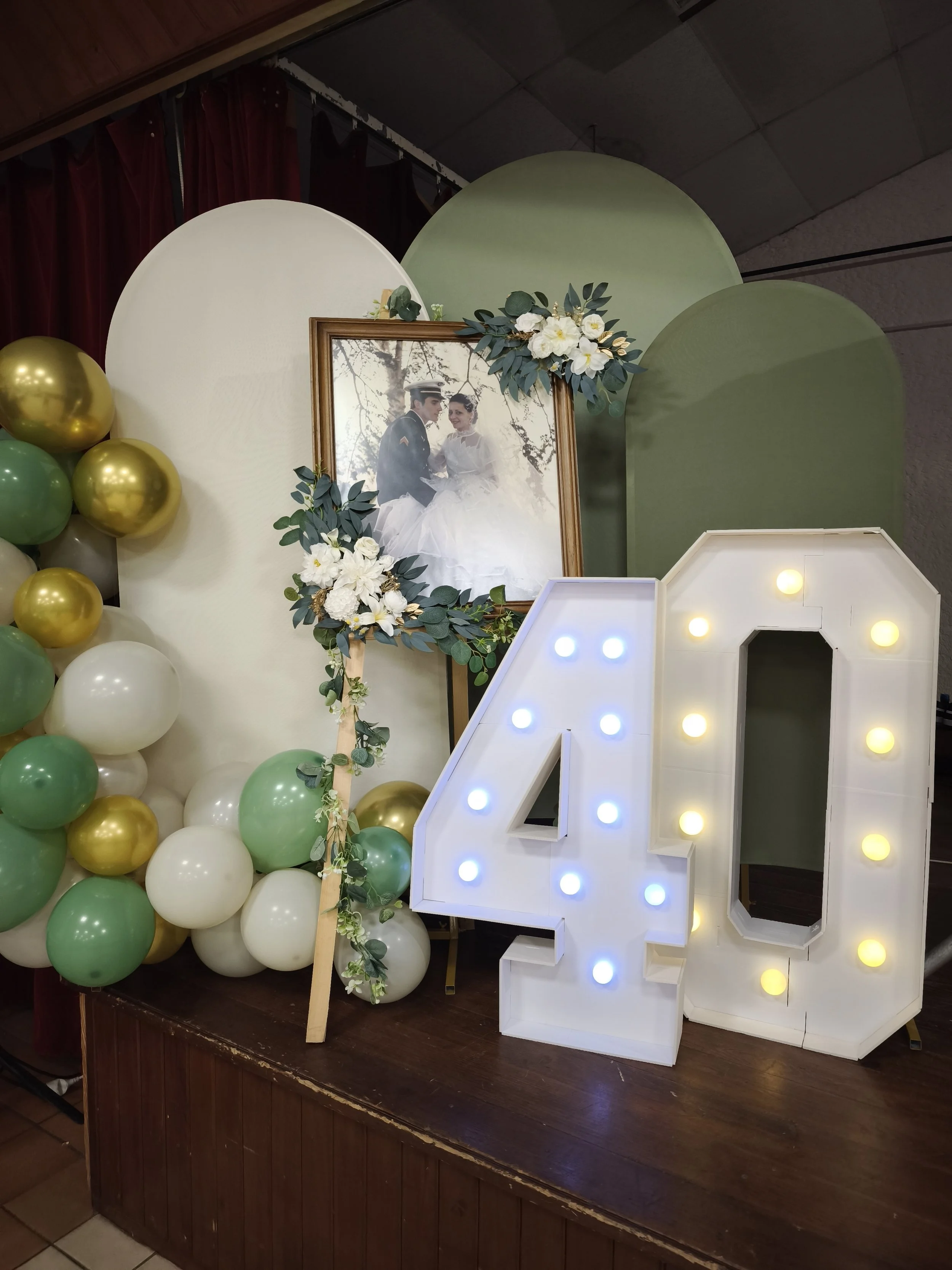 Décoration pour un événement avec un grand chiffre 4 blanc avec ampoules lumineuses, ballons verts, blancs et dorés, et un cadre photo contenant une photo de mariage.