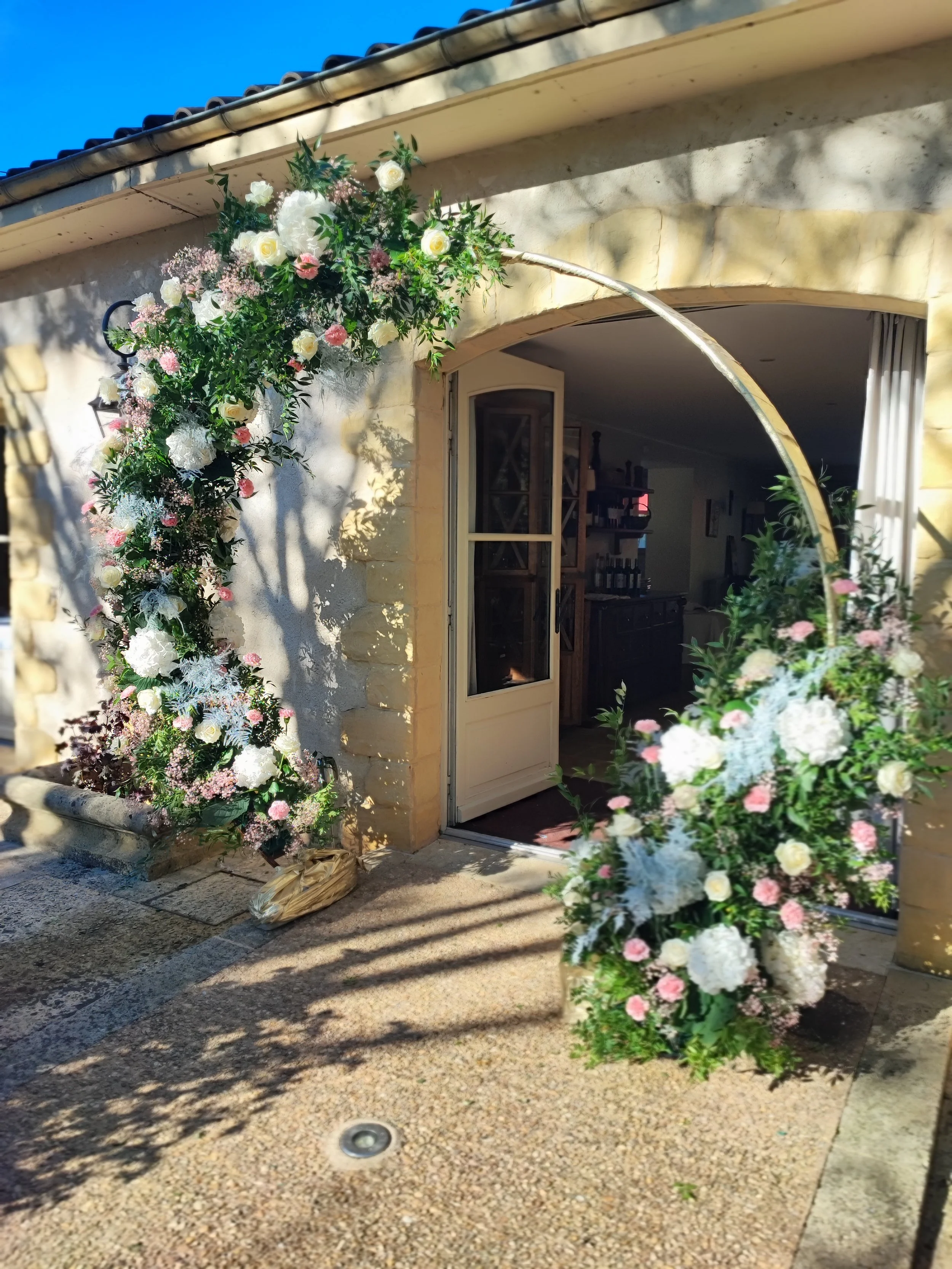 Une arche de fleurs décorative à l'entrée d'une maison en pierre avec porte ouverte, entourée de plantes et de fleurs dans un jardin en plein soleil.