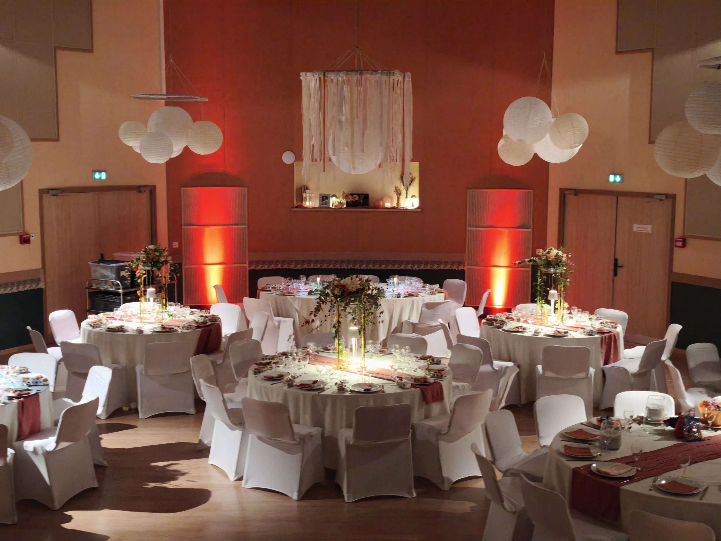 Salle de fête décorée avec tables rondes couvertes de nappes blanches, ornées de bouquets de fleurs et de vaisselle, murs avec éclairage rouge et lampions suspendus.