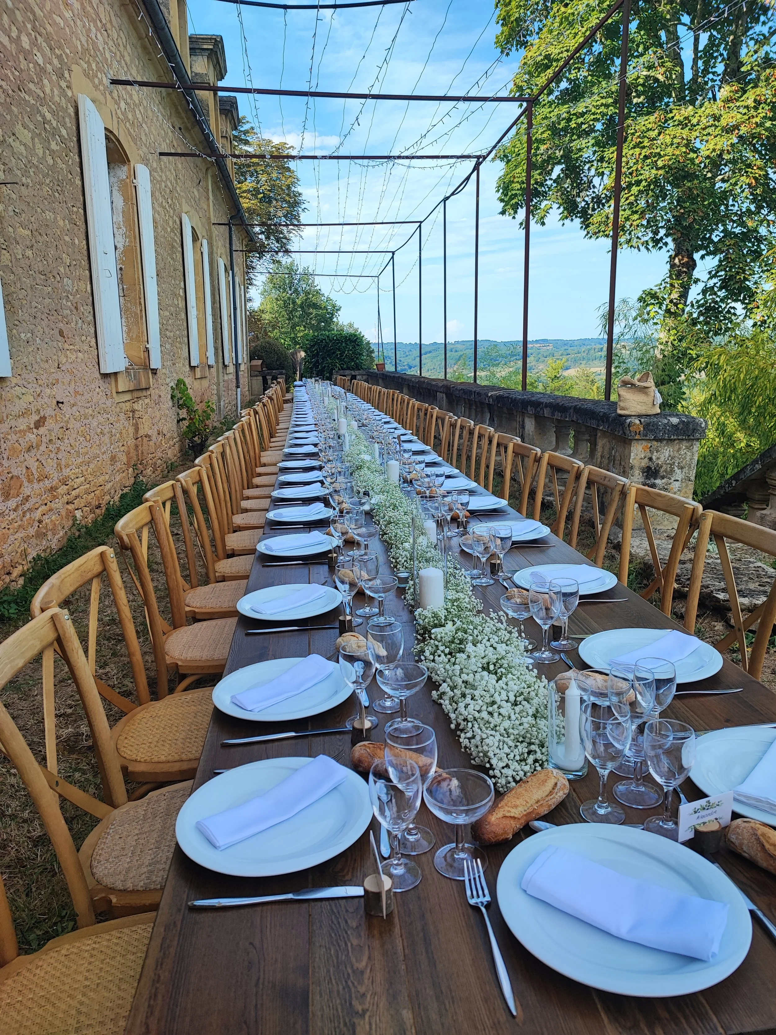 Long banquet table décoré avec des fleurs blanches, des chandelles, des assiettes blanches, des verres à vin, des serviettes blanches pliées, des baguettes et du pain, à l'extérieur avec vue sur la campagne et un ciel bleu.
