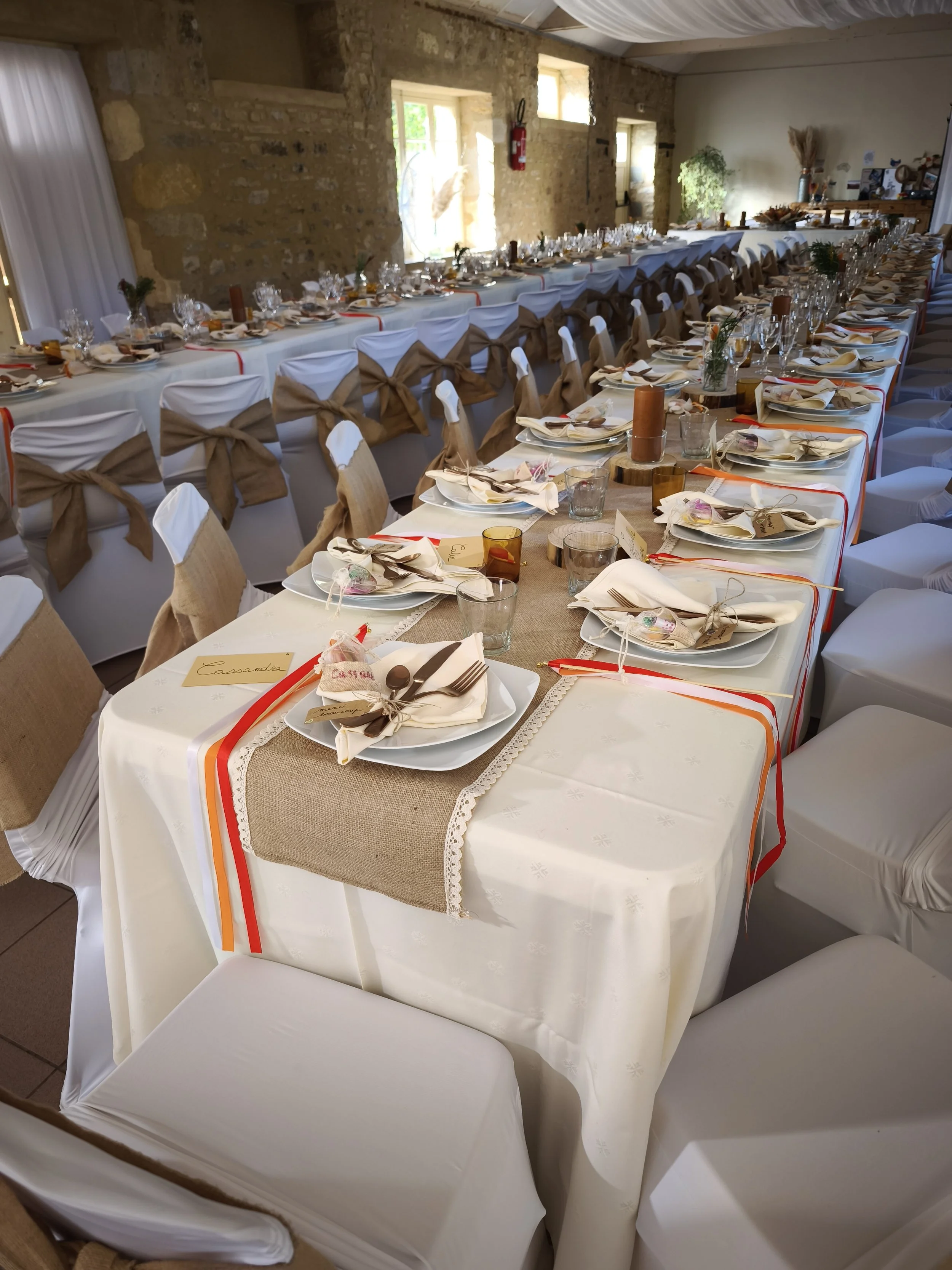 Une grande table décorée pour un événement, avec des assiettes, des verres, des couverts, des serviettes, des bougies, et des petits cadeaux, dans une salle avec des murs en pierre et des fenêtres.