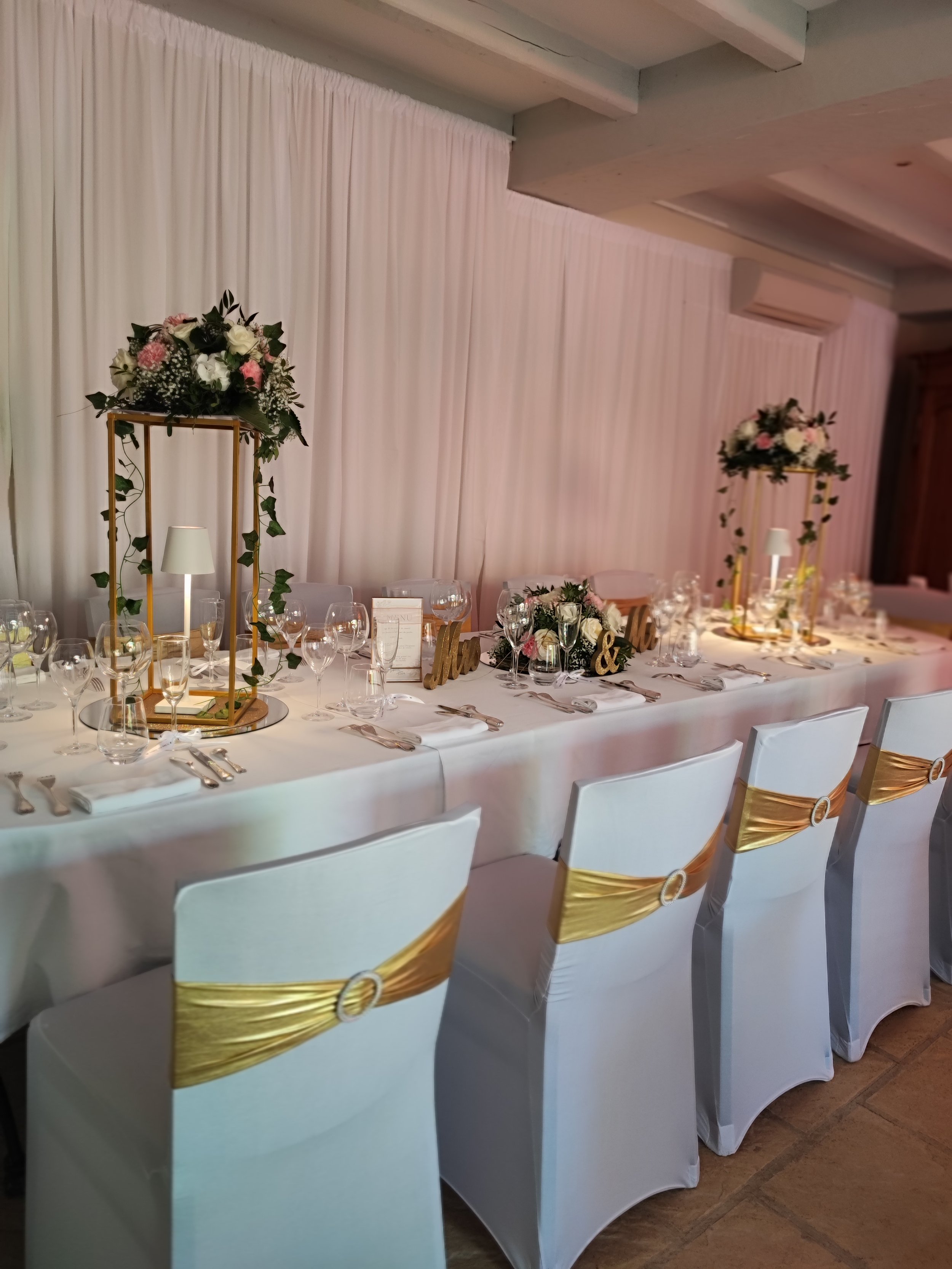 Table de mariage décorée avec des fleurs, des centres de table, des bougies et des assiettes, dans une salle ornée de rideaux blancs et d'une décoration élégante.