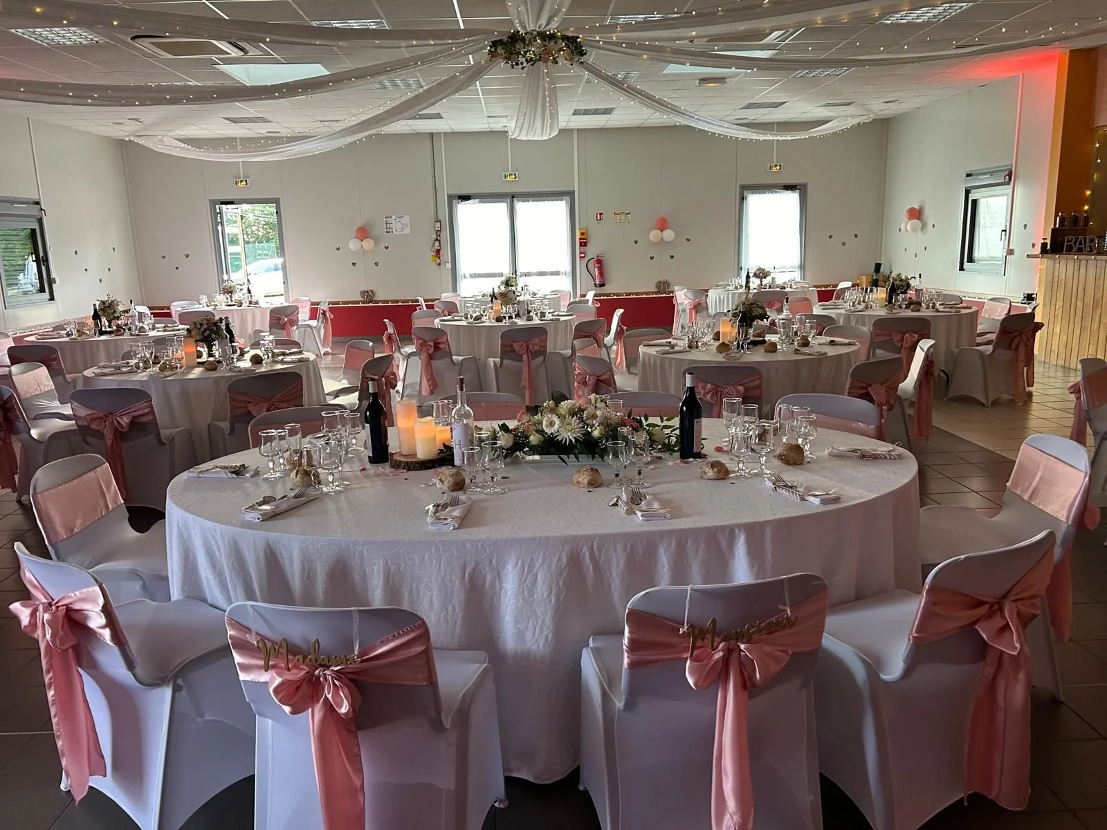 Salle de réception décorée pour un mariage avec tables rondes, nappes blanches, chaises avec rubans roses, centres de table floraux, bougies et bouteilles de vin.
