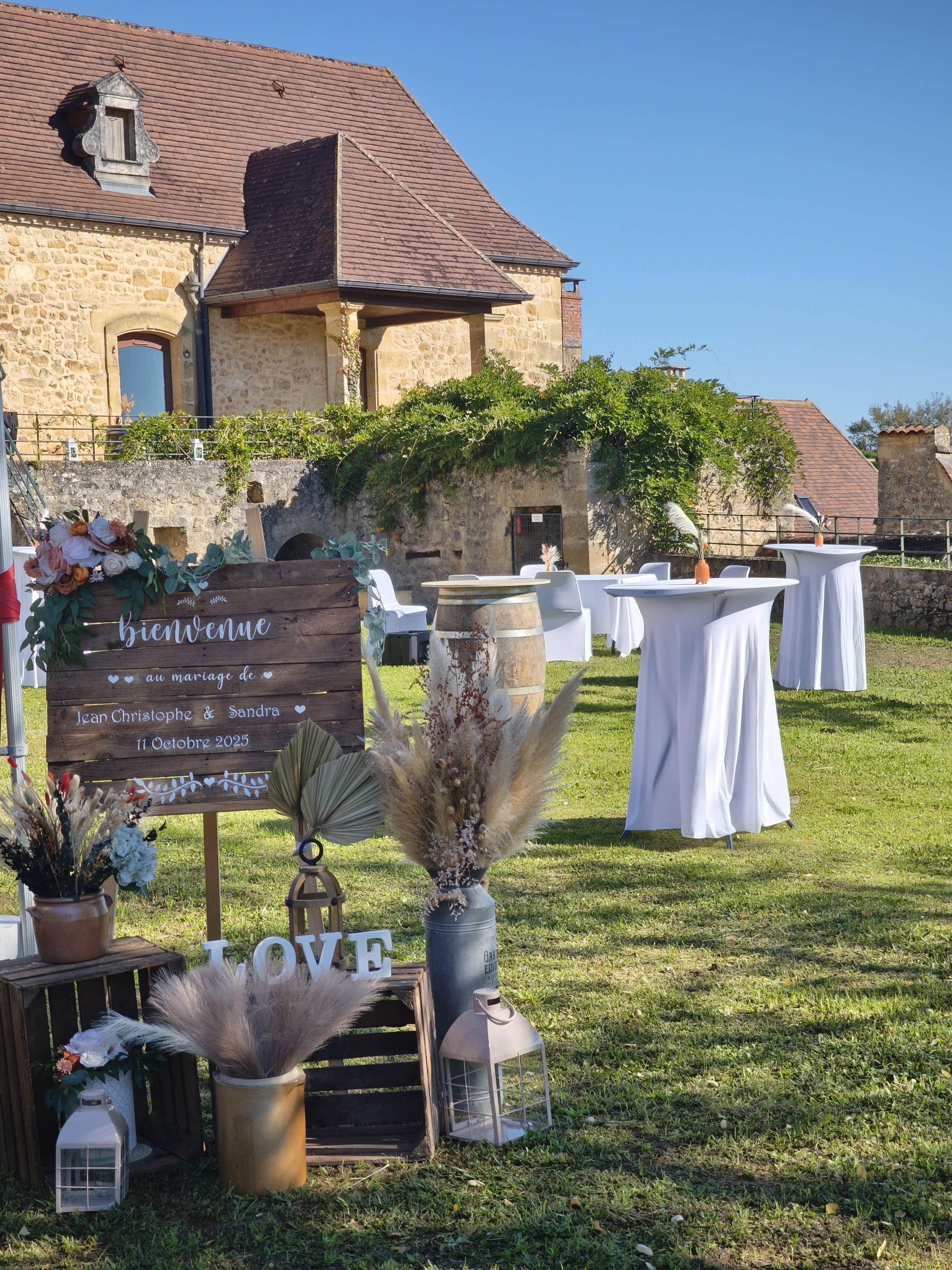 Décoration de mariage en plein air avec tables rondes drapées de blanc, une enseigne en bois avec écrit 'bienvenue au mariage de Jean Christophe & Sandra, 11 Octobre 2025', fleurs, lanternes et éléments en bois, dans un jardin avec une ancienne maiso