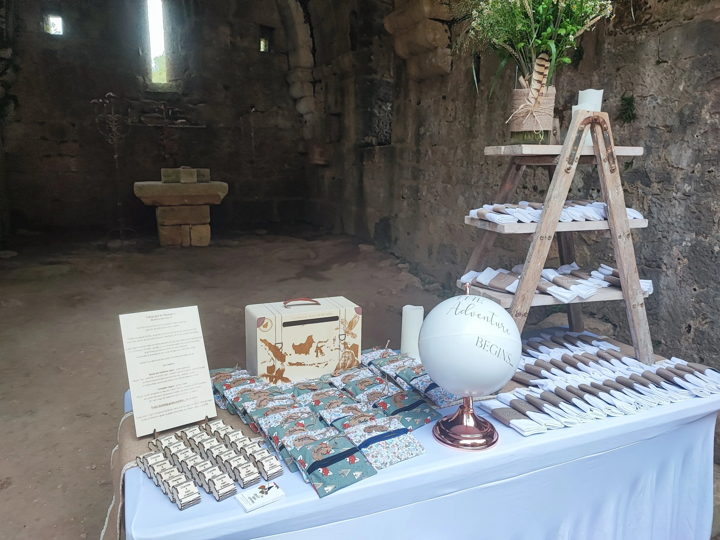Une table et un présentoir avec des articles de mariage ou de fête, dont des bibelots, des sachets et des éléments décoratifs, dans un lieu ancien avec murs en pierre.