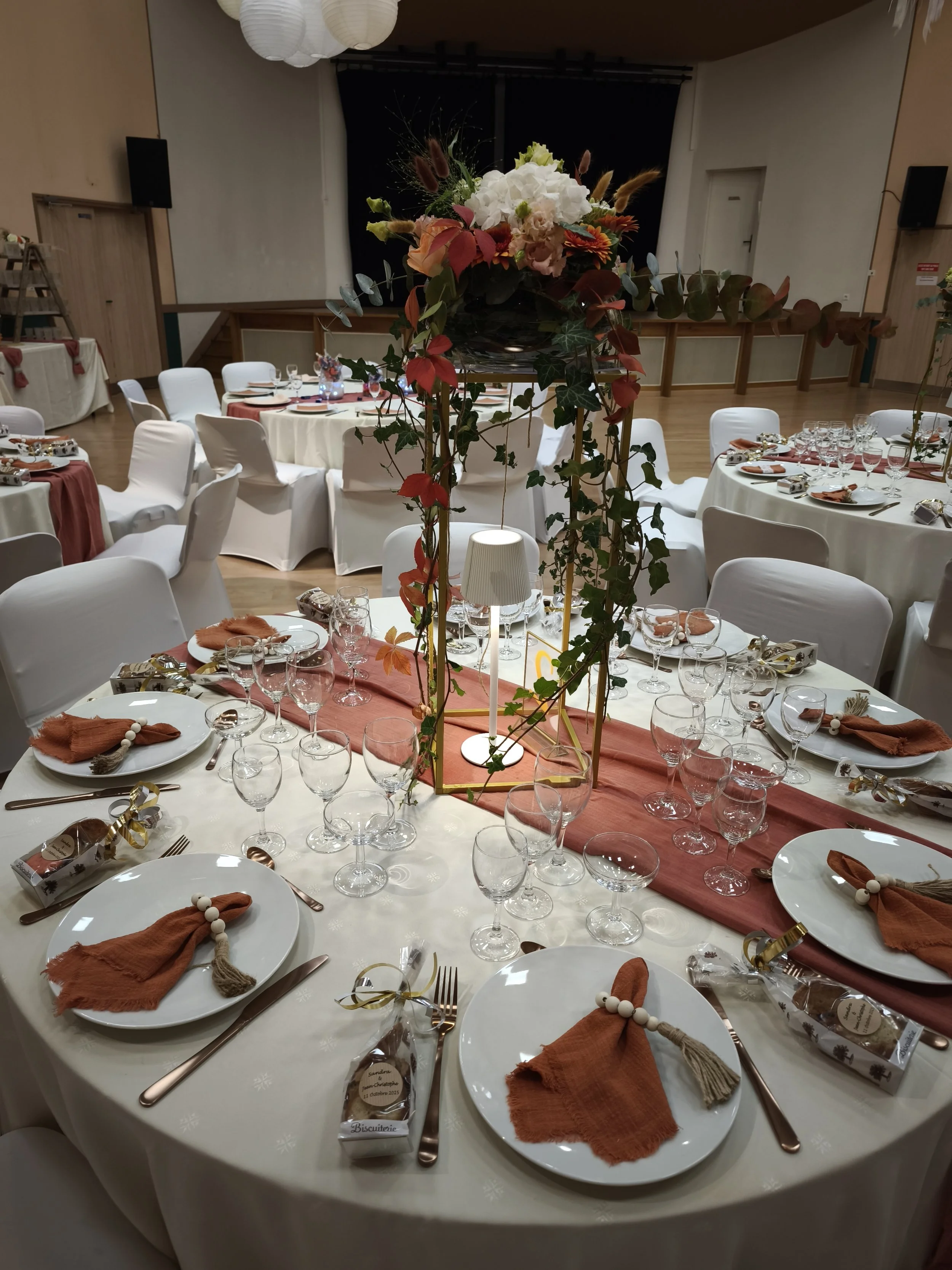 Table de fête élégamment décorée avec des assiettes, couverts, verres, serviettes en tissu orange, et un centre de table avec des fleurs. Ambiance festive dans une salle de réception.