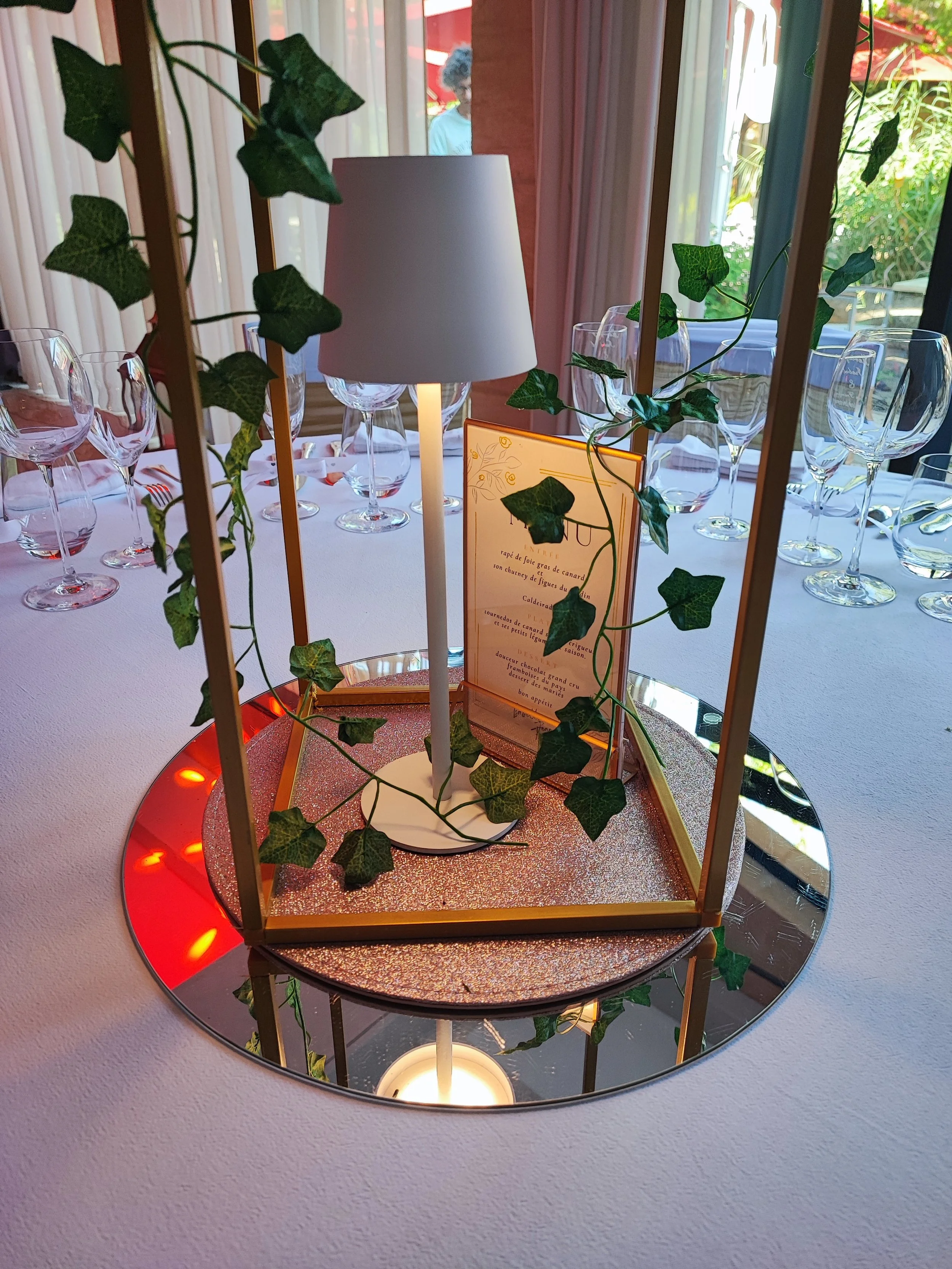 Une lampe de bureau blanche placée au centre d'une décoration verte avec des feuilles artificielles, entourée d'un miroir rond. Un menu est visible derrière la lampe, avec du texte en français. La scène est prise à l'intérieur, avec une fenêtre en ar
