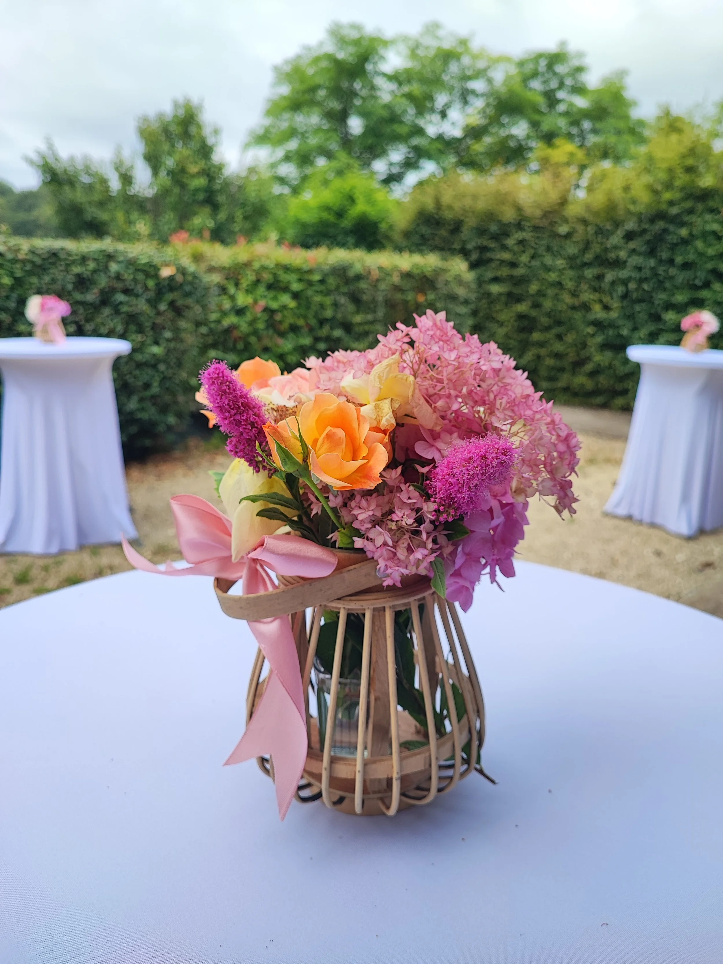 Un bouquet de fleurs colorées dans un panier en rotin placé sur une table blanche en extérieur, avec un fond de haie et d'arbres verdoyants.