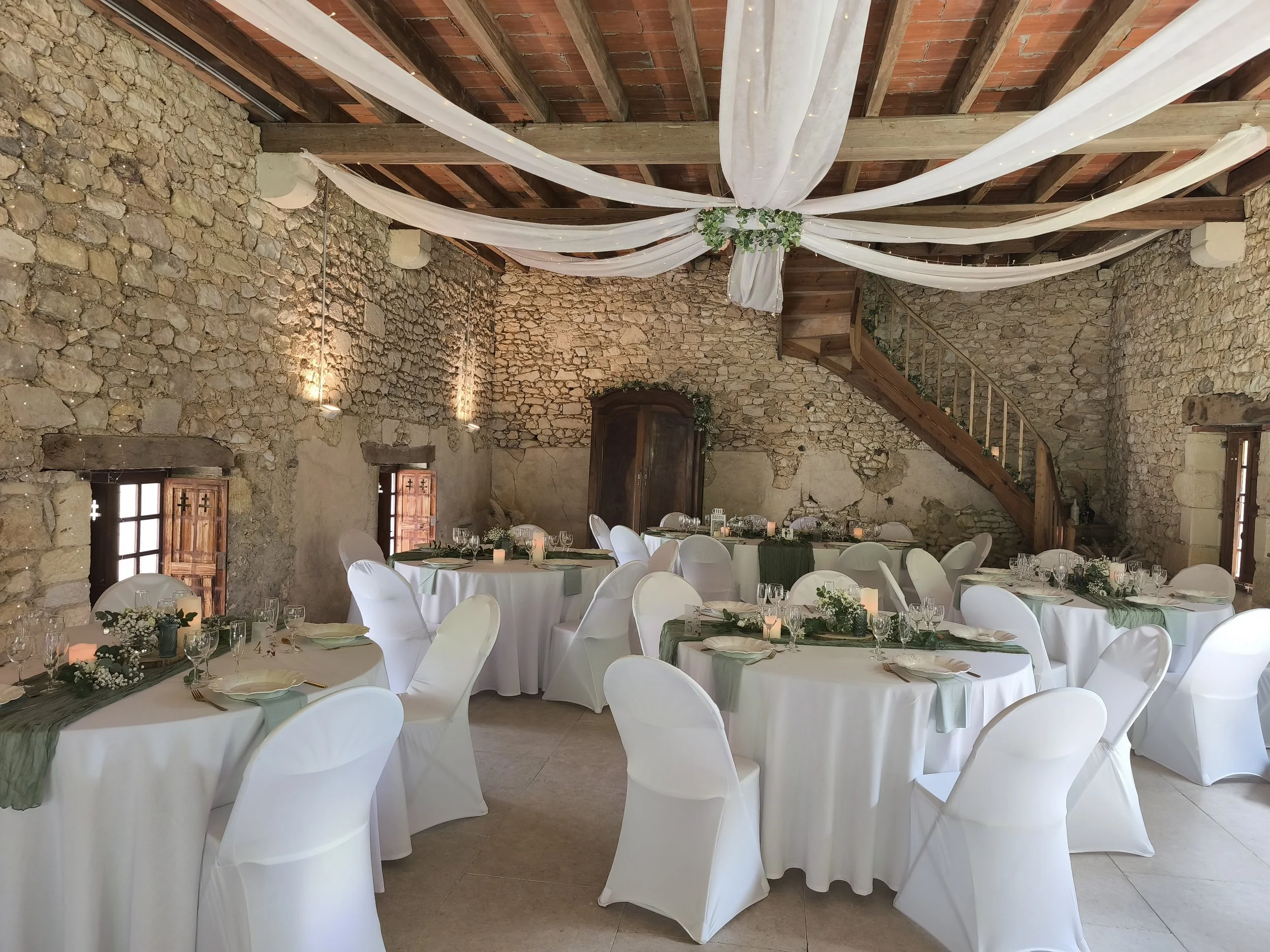 Salle de réception décorée avec des tables rondes, des chaises blanches, des bougies et des arrangements floraux, dans un bâtiment en pierre avec poutres en bois et draperies blanches suspendues au plafond.