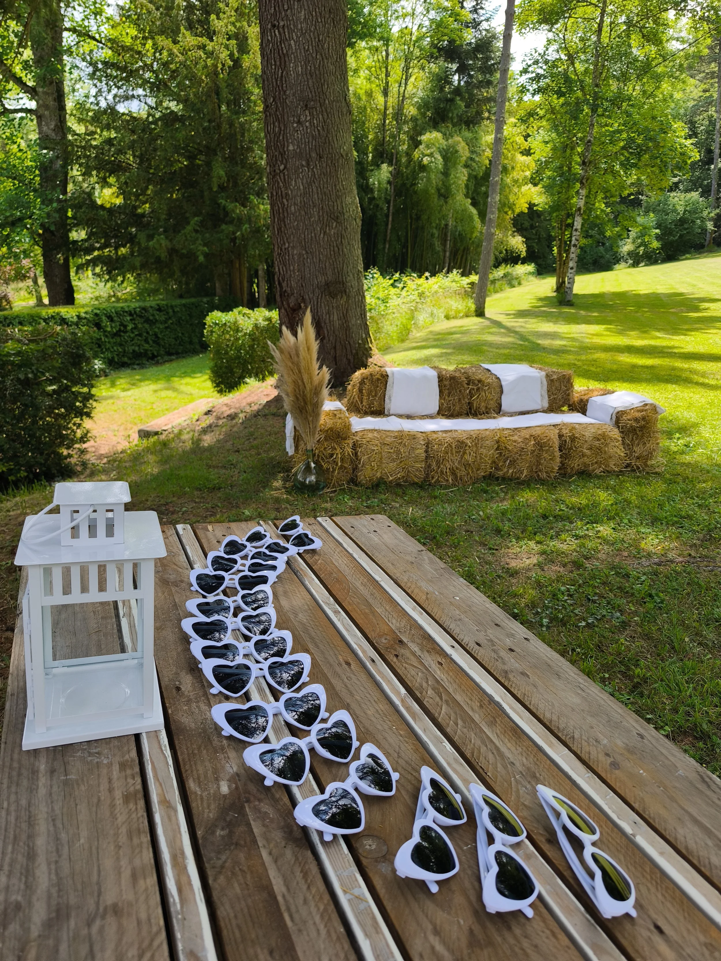 L'image montre une table en bois avec plusieurs paires de lunettes de soleil blanches en forme de cœur, ainsi qu'une petite lanterne blanche et un vase avec des fleurs. En arrière-plan, il y a une terrasse en plein air au milieu d'un jardin verdoyant