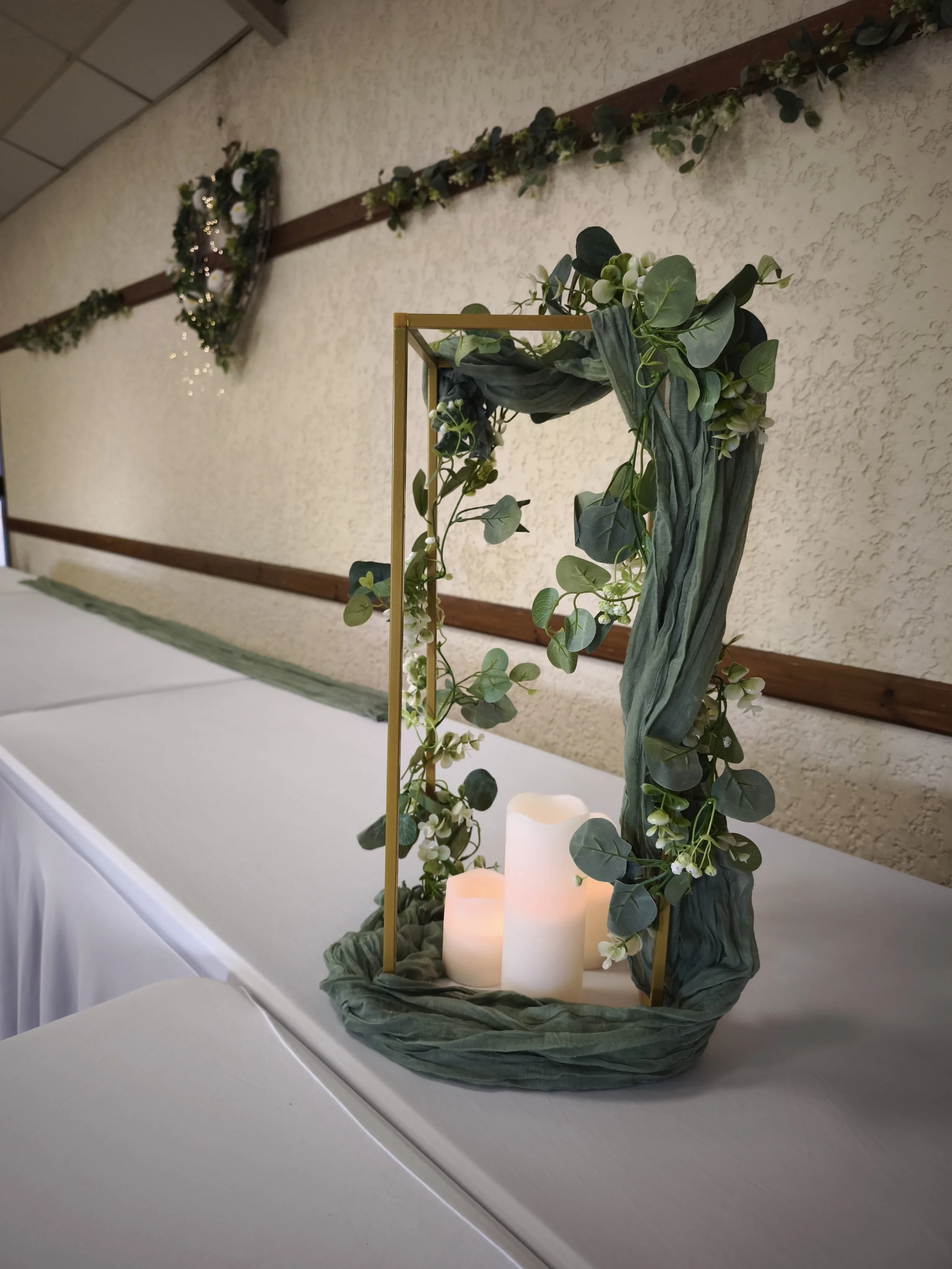 Décoration florale avec bougies sur une table pour un événement, comprenant des feuilles vertes et des fleurs blanches, dans un espace intérieur.
