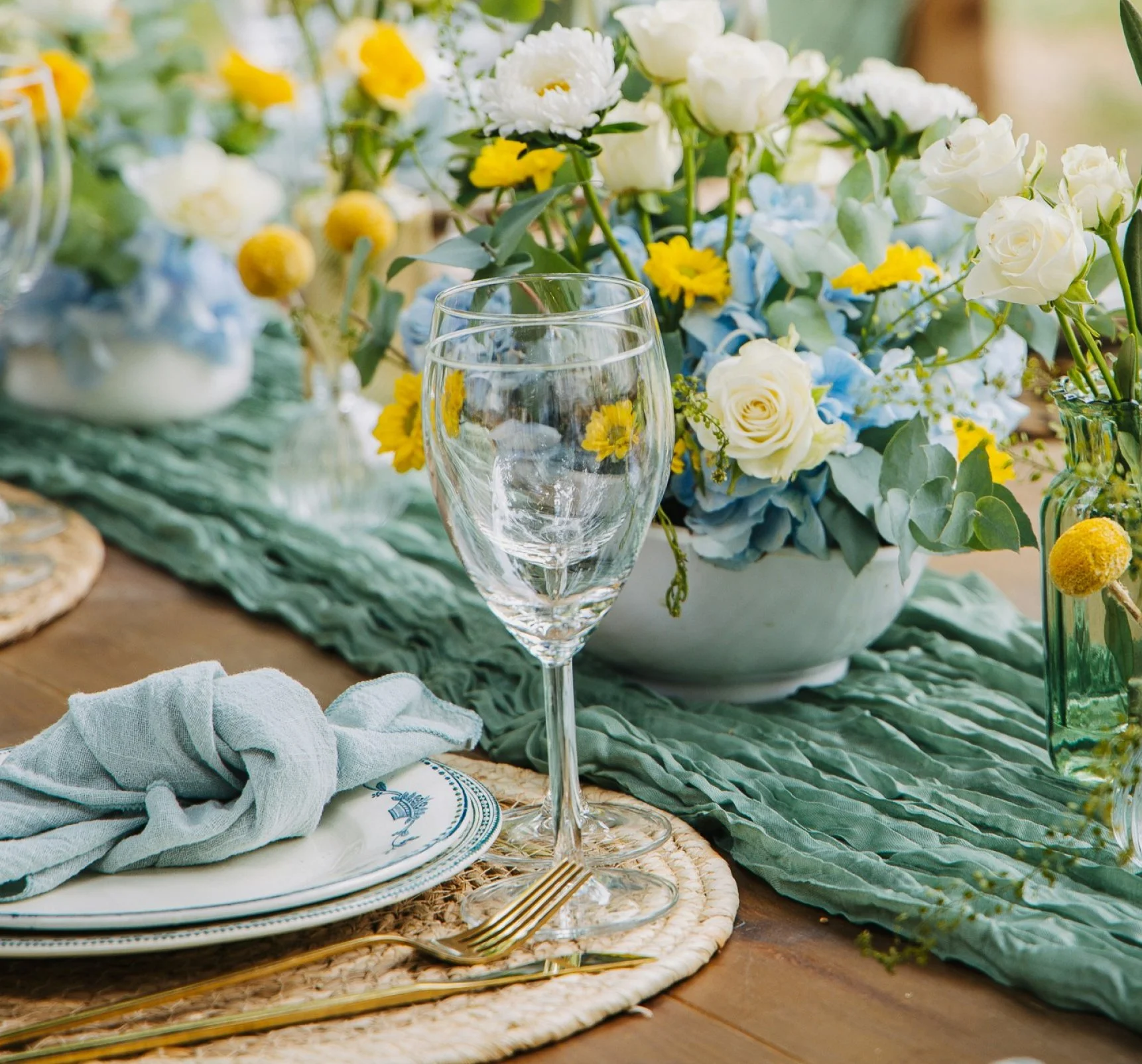 Une table décorée avec des fleurs blanches, jaunes et bleues, entourée de vaisselle élégante, un verre à vin et des couverts en or. Tissu bleu clair, nappe en raphia, ambiance festive et raffinée.