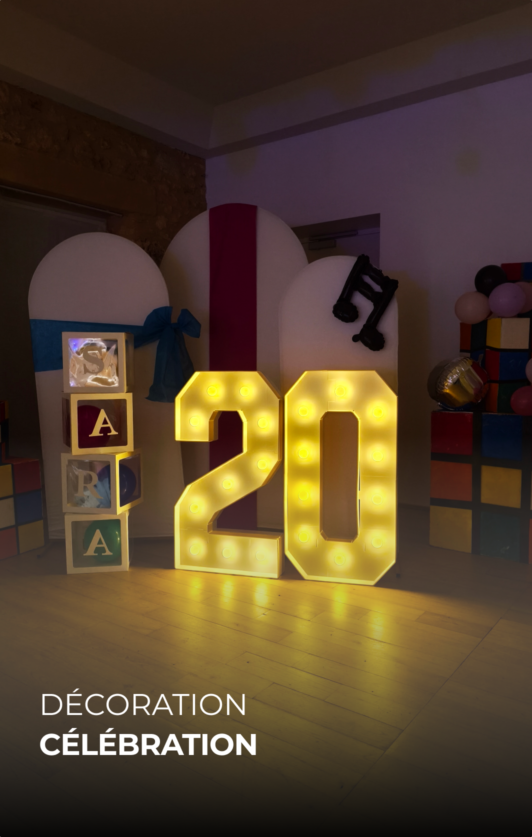 Décoration de fête avec un grand chiffre lumineux '20', cubes avec lettres 'S', 'A', 'R', 'A', et un décor coloré en arrière-plan.