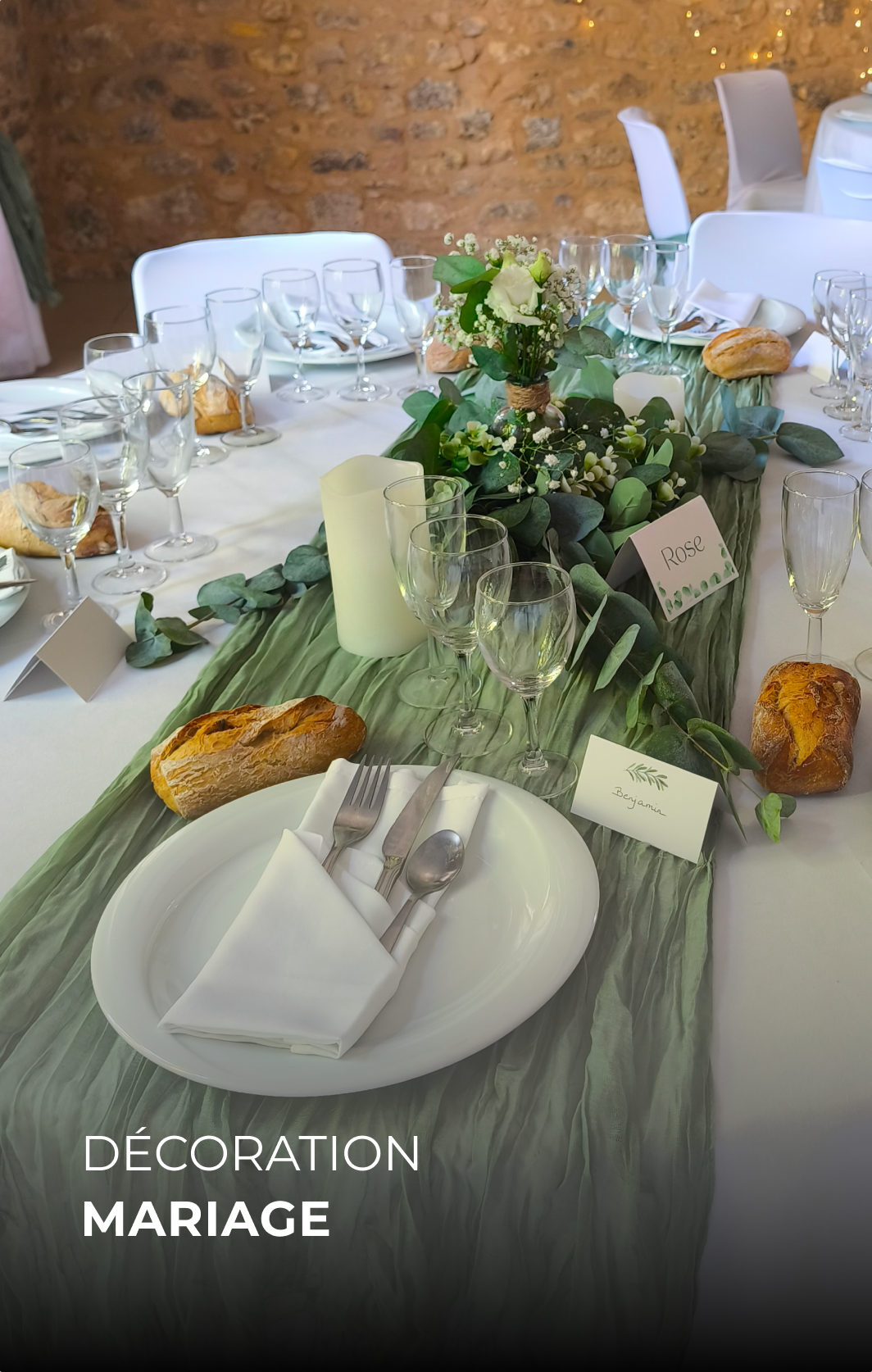 Table de mariage décorée avec des fleurs, des pains, des verres, des assiettes, des couverts, et des cartes de noms, avec un chemin de table en tissu vert et un fond en briques