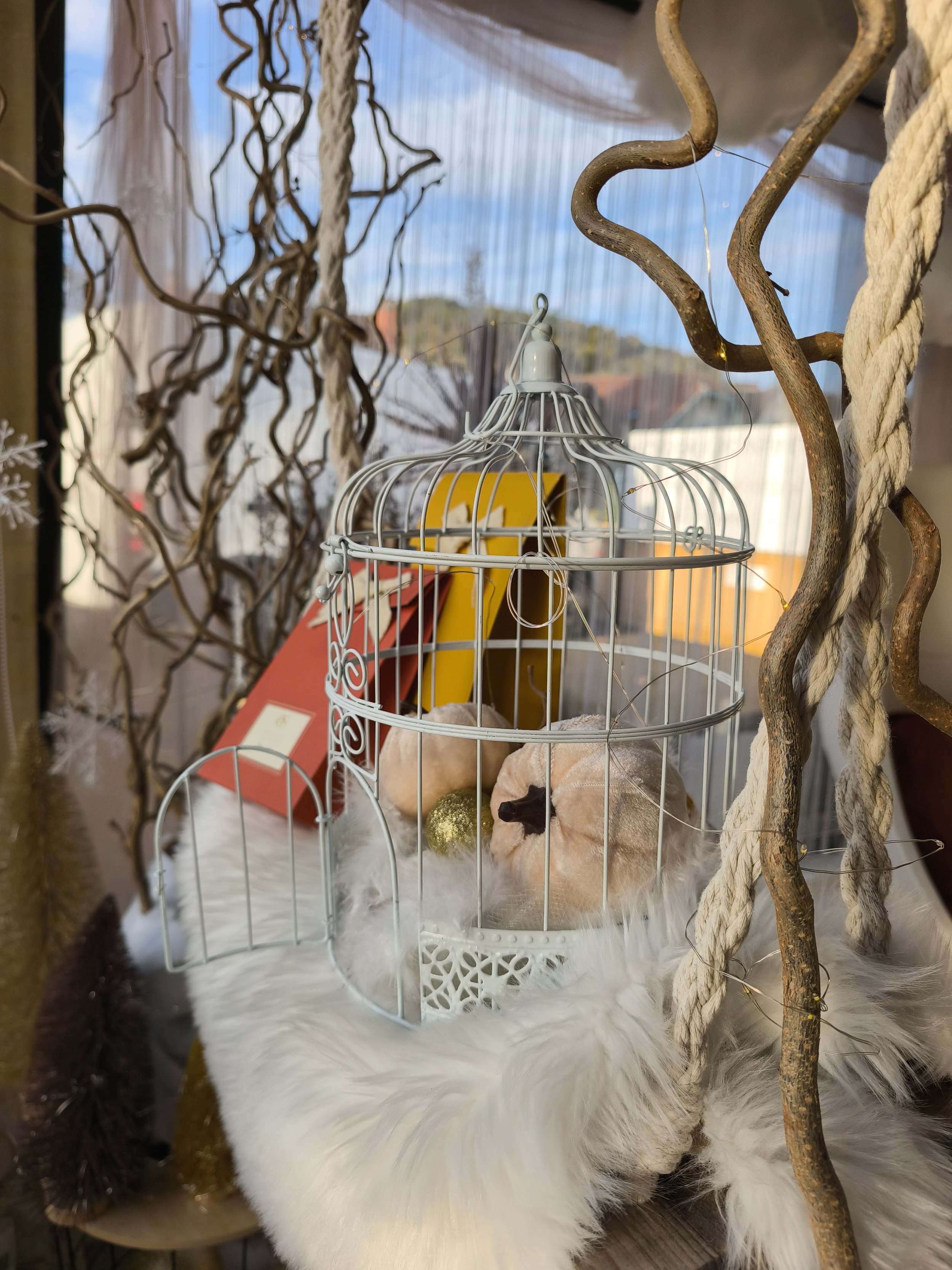 Une décoration de Noël avec un petit animal en peluche, une cage blanche, des boules dorées, et des branches décoratives sur un fond de fenêtre avec un ciel bleu