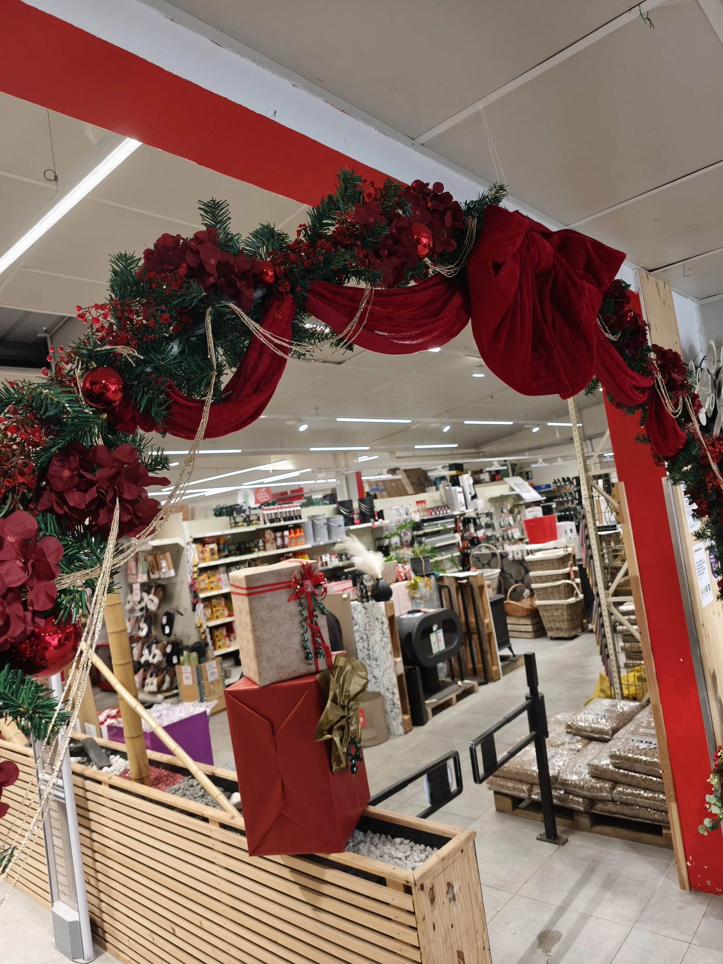 Décoration de Noël avec guirlande de plantes vertes, fleurs rouges, boules rouges et rubans rouges dans un magasin, avec des étagères remplis d'articles et des cadeaux emballés en arrière-plan.