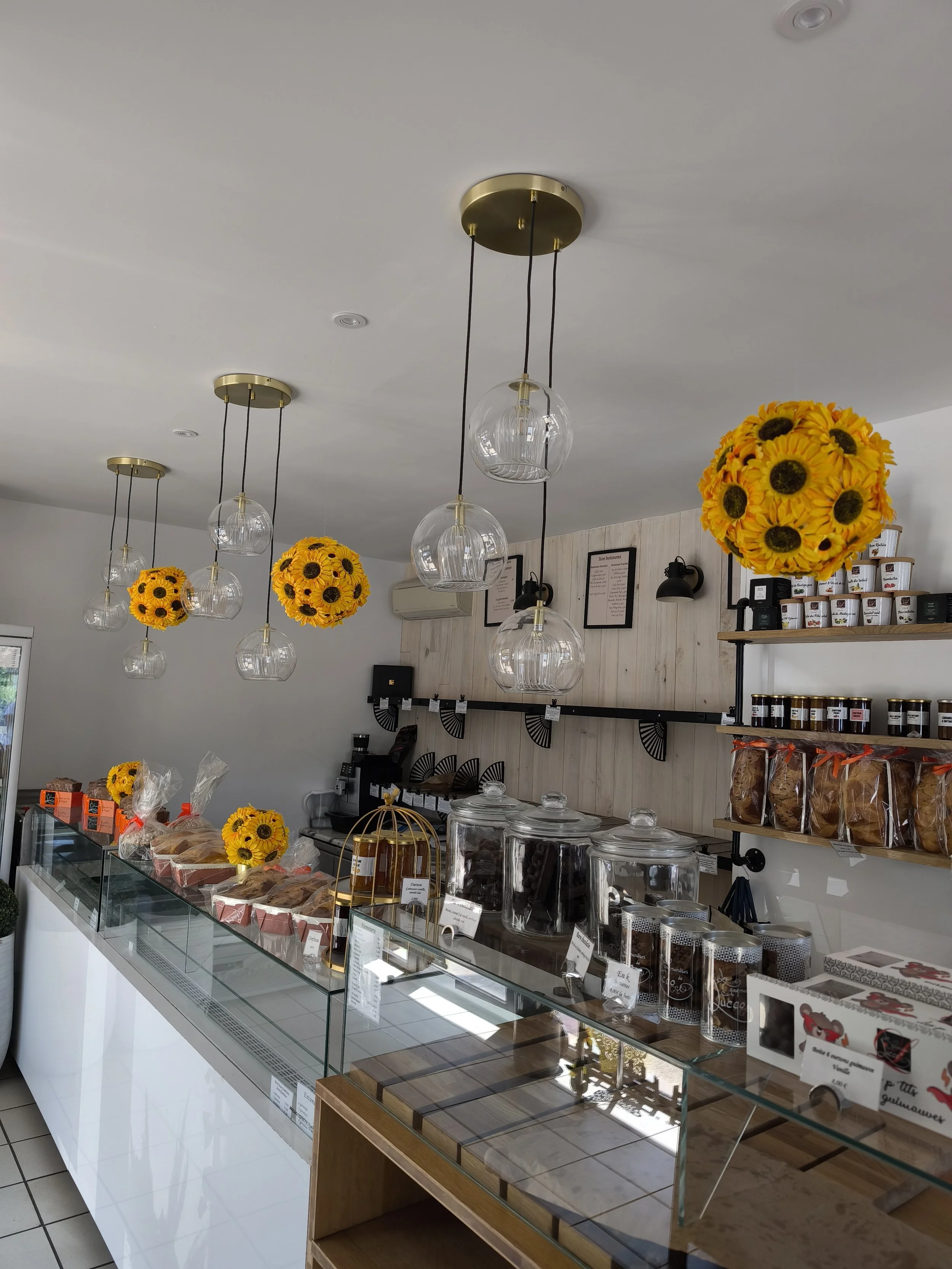 Vitrine d'une boutique de pâtisserie avec plusieurs présentations de gâteaux, biscuits et confiseries, décorée avec des bouquets de tournesols suspendus au plafond.
