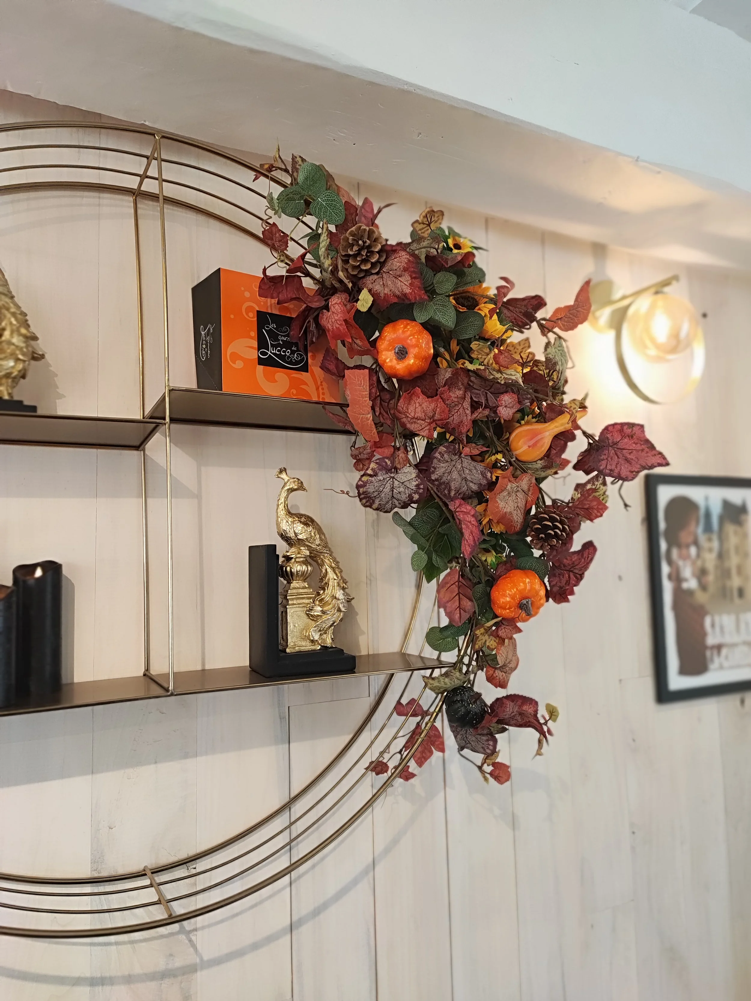 Décoration intérieure avec une étagère en métal doré, contenant un pot orange avec une décoration végétale aux feuilles vertes et rouges, des pommes de pin et une petite sculpture d'un animal doré.