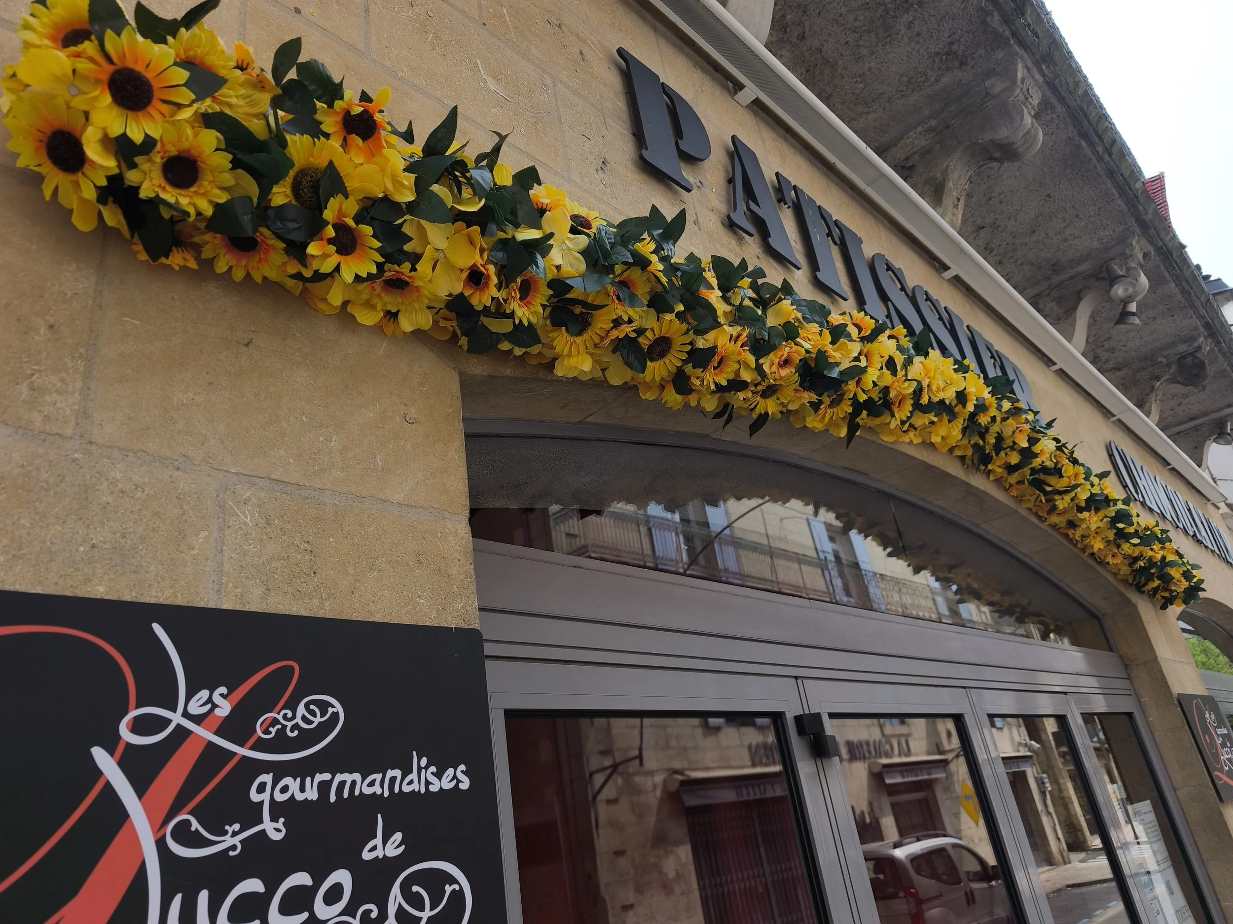 Une façade de bâtiment avec une décoration florale composée de fleurs jaunes et de feuilles vertes, ainsi qu'un panneau noir avec des écritures en cursive blanche. Au-dessus, il y a des lumières et en bas, une partie d'un véhicule est visible.