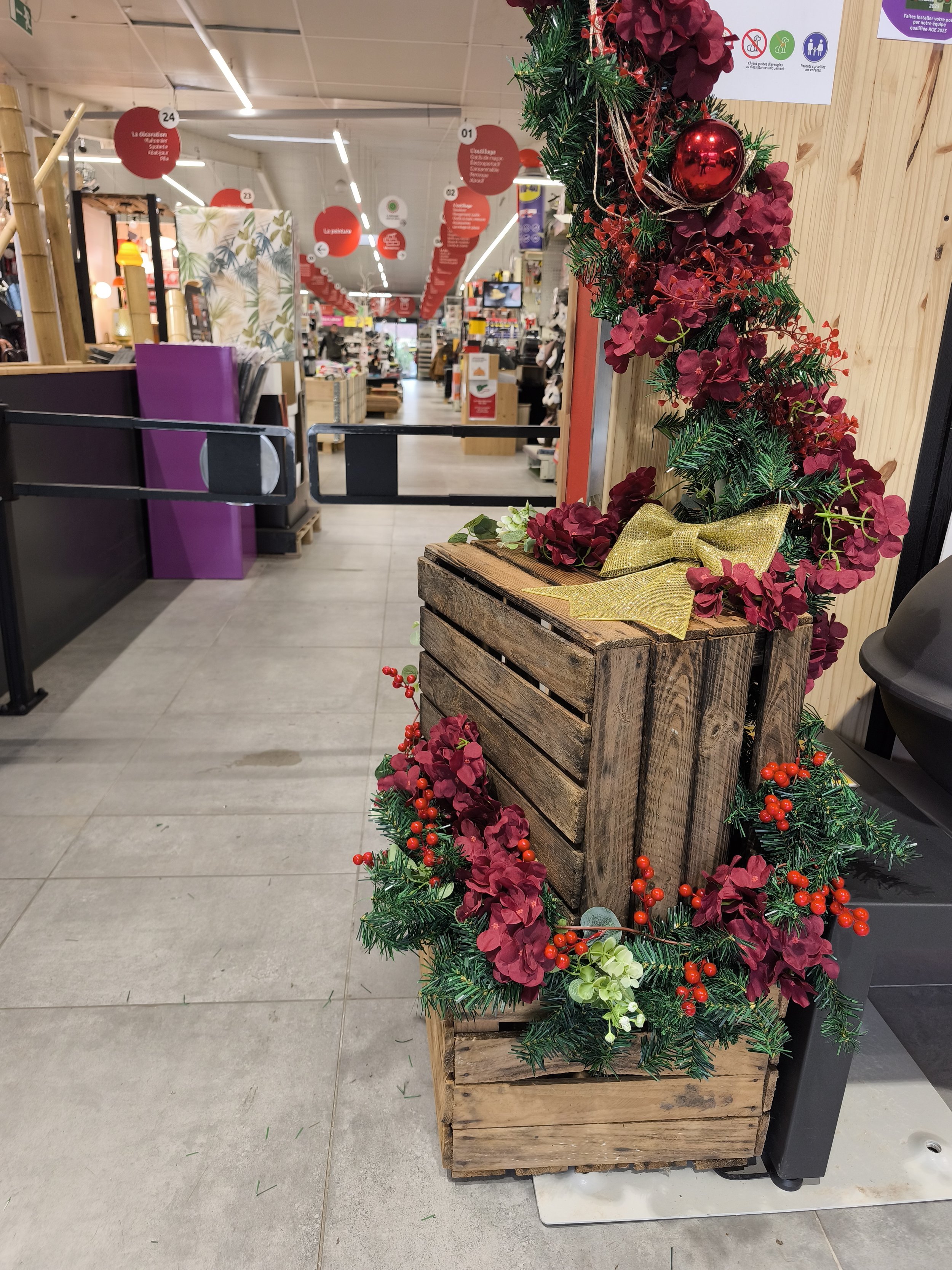 Décoration de Noël faite de branches d'épicéas, de boules rouges, de fleurs rouges et blanches, avec un gros nœud doré, posée sur une caisse en bois.