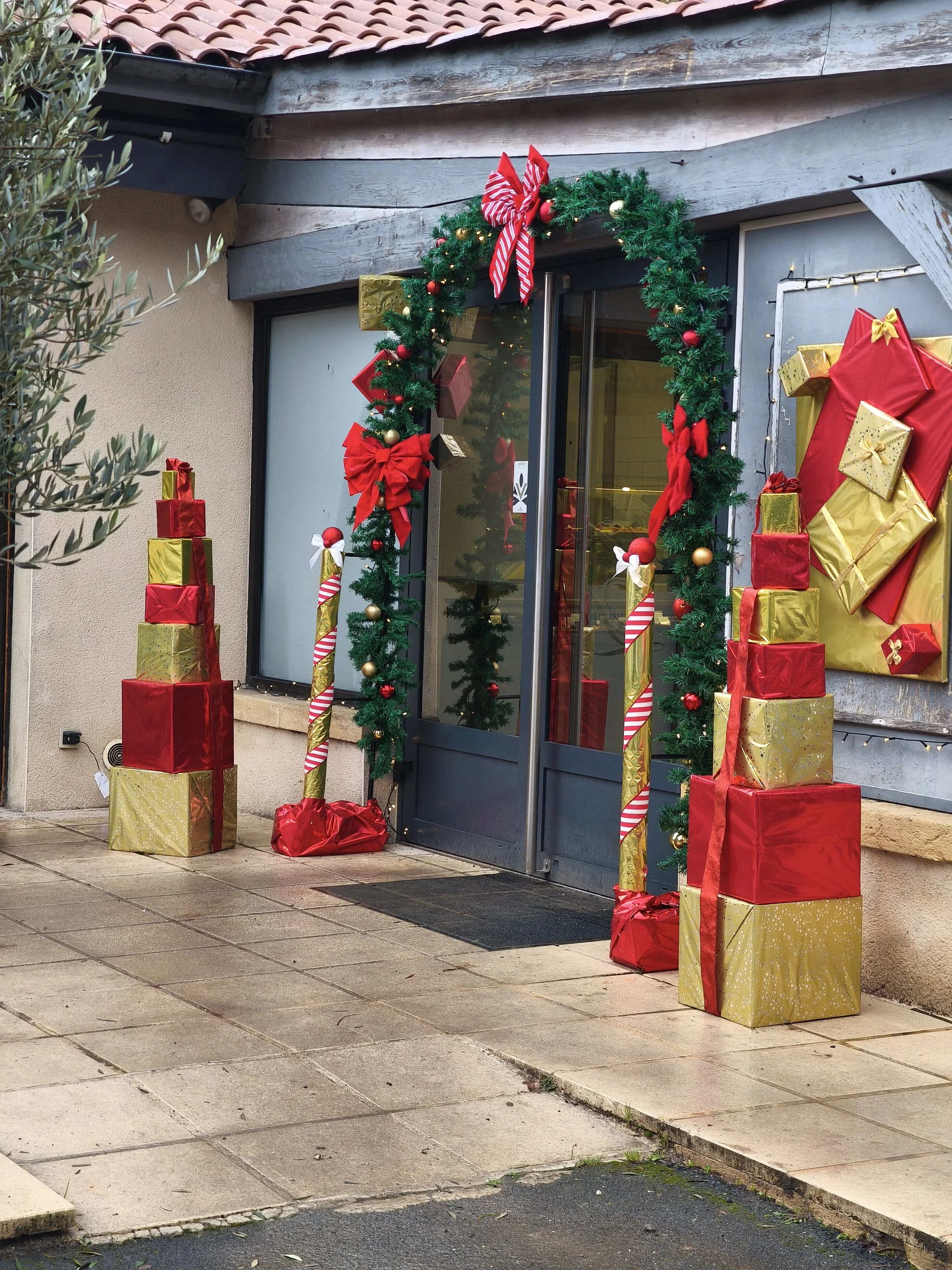 Décoration de Noël à l'extérieur d'une maison avec une porte d'entrée encadrée de guirlandes vertes, de rubans rouges et or, de boules de Noël rouges et or, ainsi que des cadeaux enveloppés en rouge et or empilés de chaque côté de la porte.