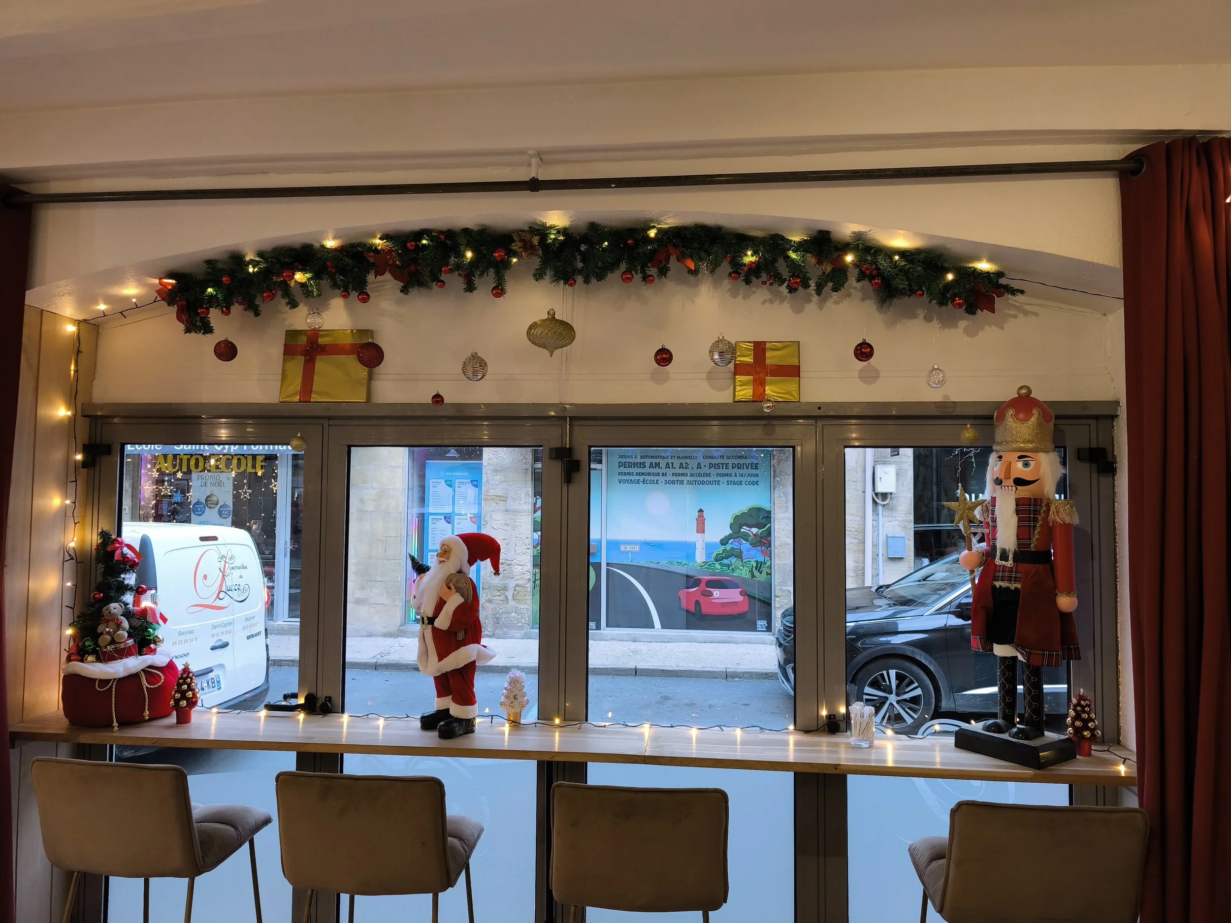 Décoration de Noël à l'intérieur d'un magasin avec une guirlande de sapin, des boules rouges et or, et des jouets de Père Noël et d'elfe. Vue depuis une table avec quatre chaises.