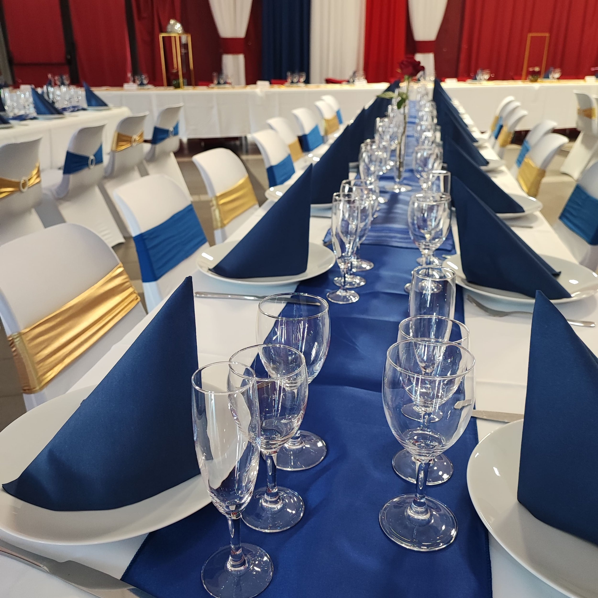 Une table de fête élégamment dressée avec des verres à vin transparents alignés, des nappes bleues foncées pliées en forme de pyramide, des chaises blanches ornées de rubans dorés, et un décor de rideaux rouges avec des accents blancs et bleus en arr