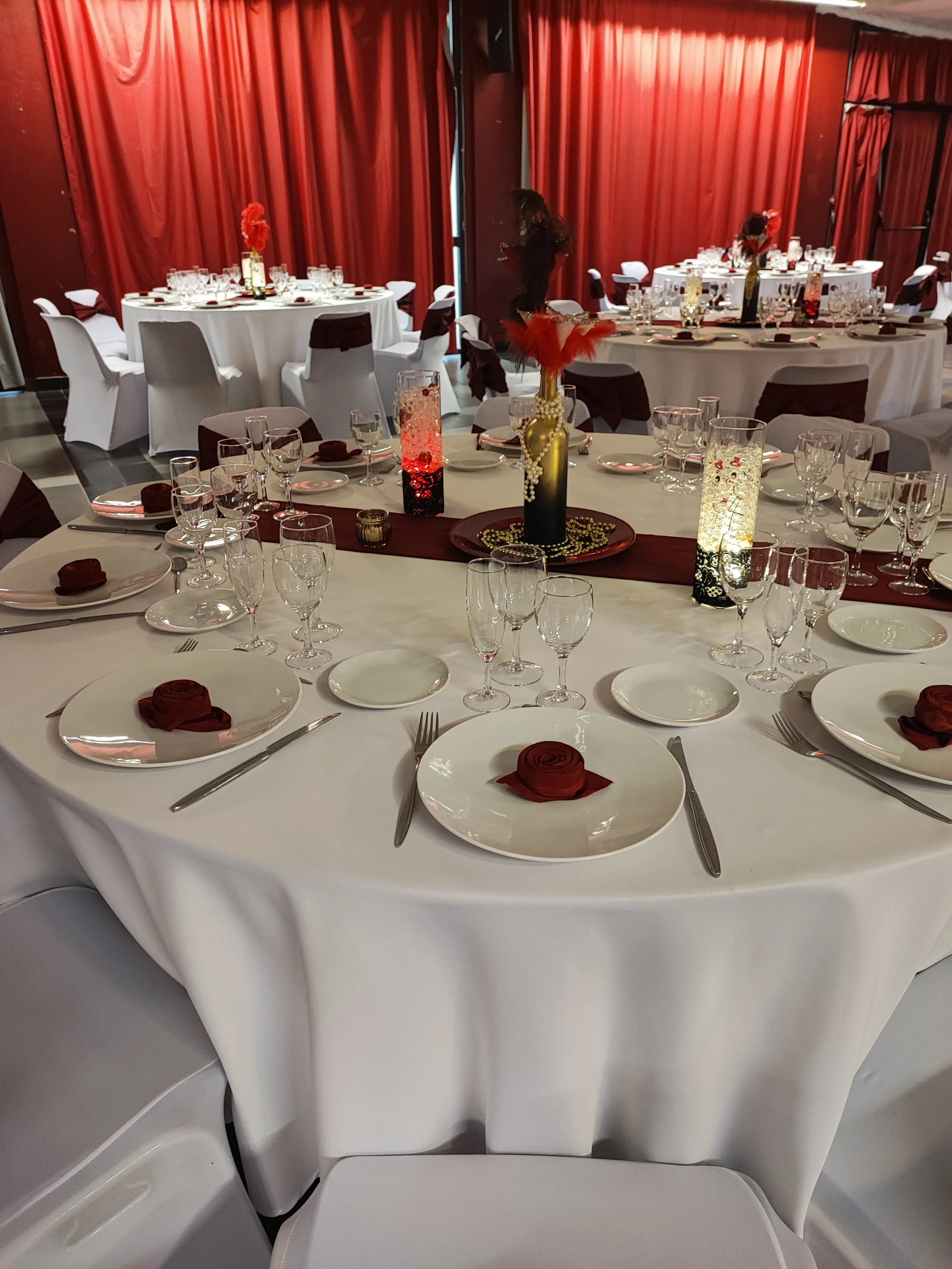 Salle de banquet élégante avec des tables rondes couvertes de nappes blanches, ornées de décorations florales rouges, de vases lumineux, et disposant de vaisselle raffinée, de serviettes enroulées en forme de rose, et de verres à vin et à eau. Le fon