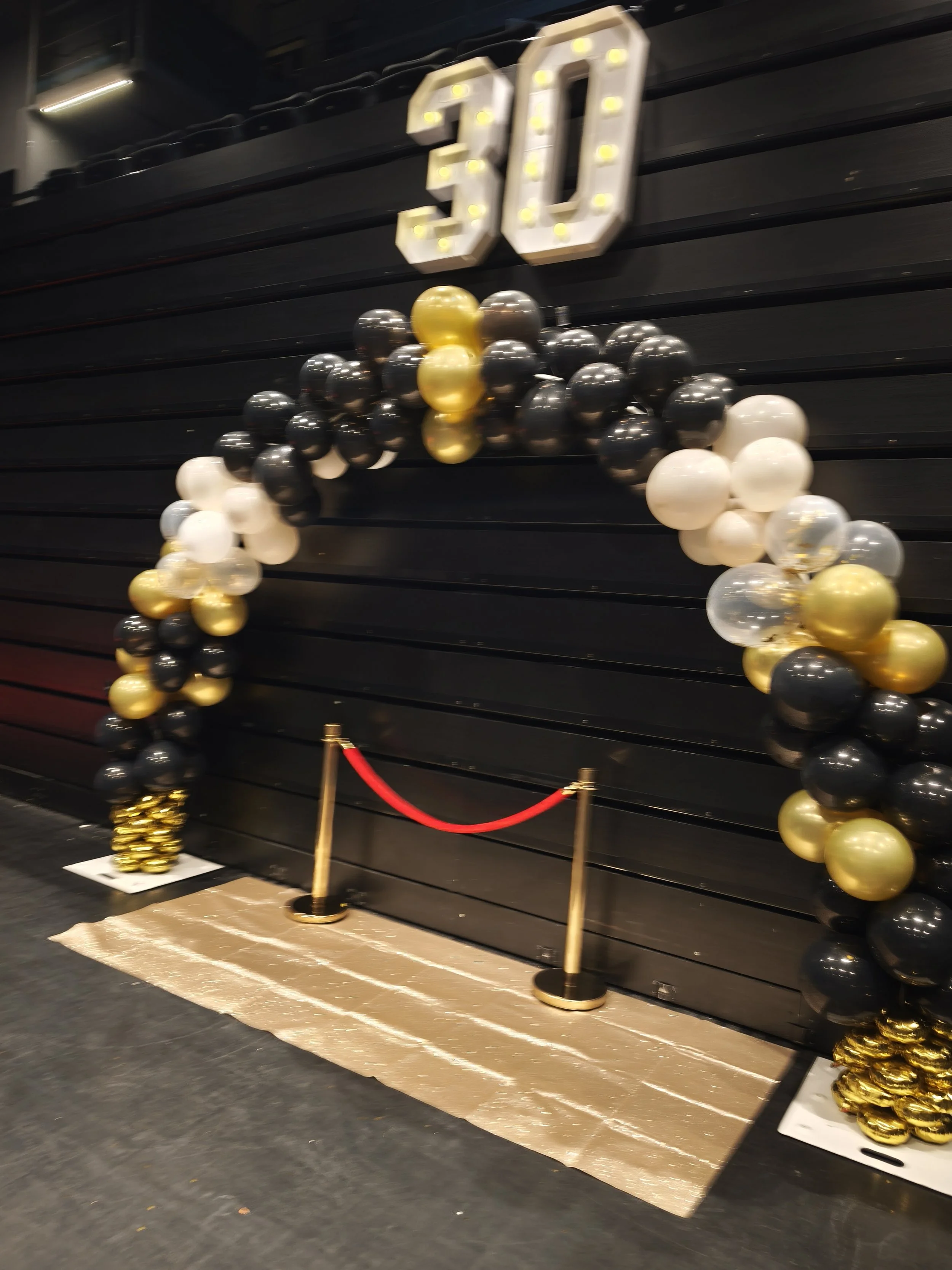 Décoration de bal du 30ème anniversaire avec un grand numéro 30 en lettres lumineuses, un arc de ballons noirs, blancs et dorés, une corde rouge pour délimiter la zone, et un tapis doré au sol.
