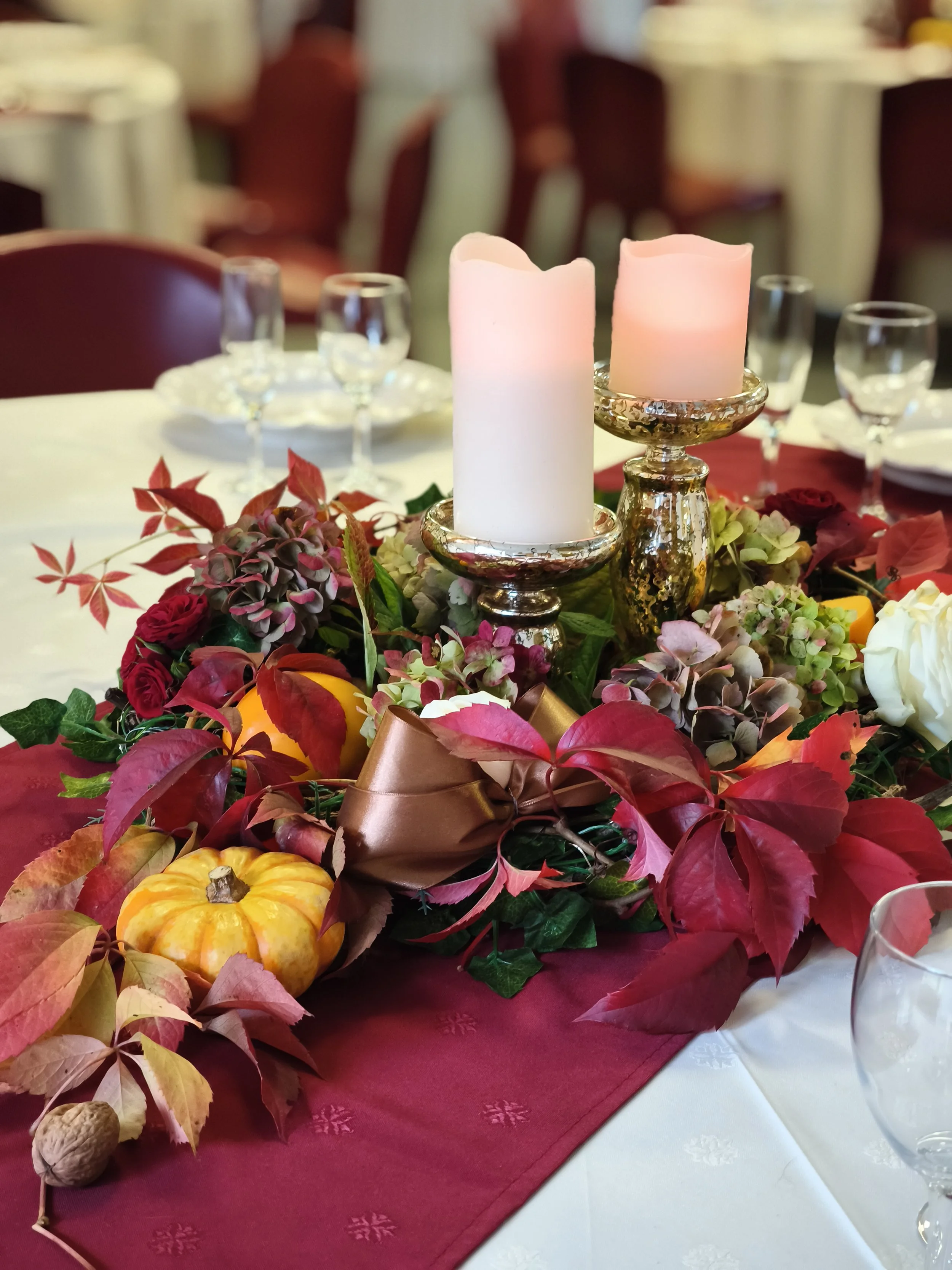 Table de fête décorée avec des bougies en forme de chandelles, entourée de fleurs colorées et de petites citrouilles, sur une nappe rouge et blanche.