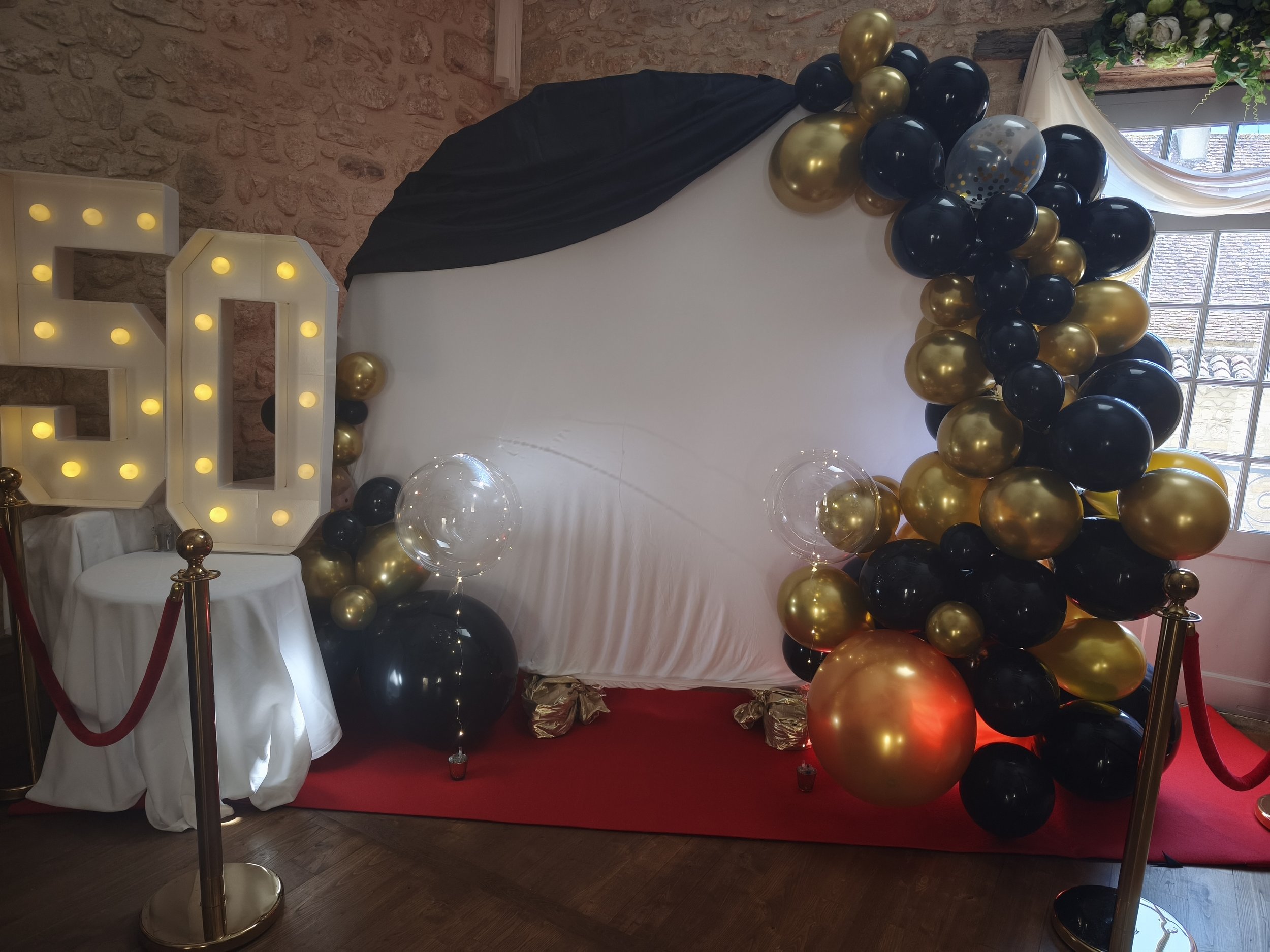 Décoration de fête avec un mur de ballons noirs et dorés, une grande lettre en lumière, et un tapis rouge, près d'une fenêtre.
