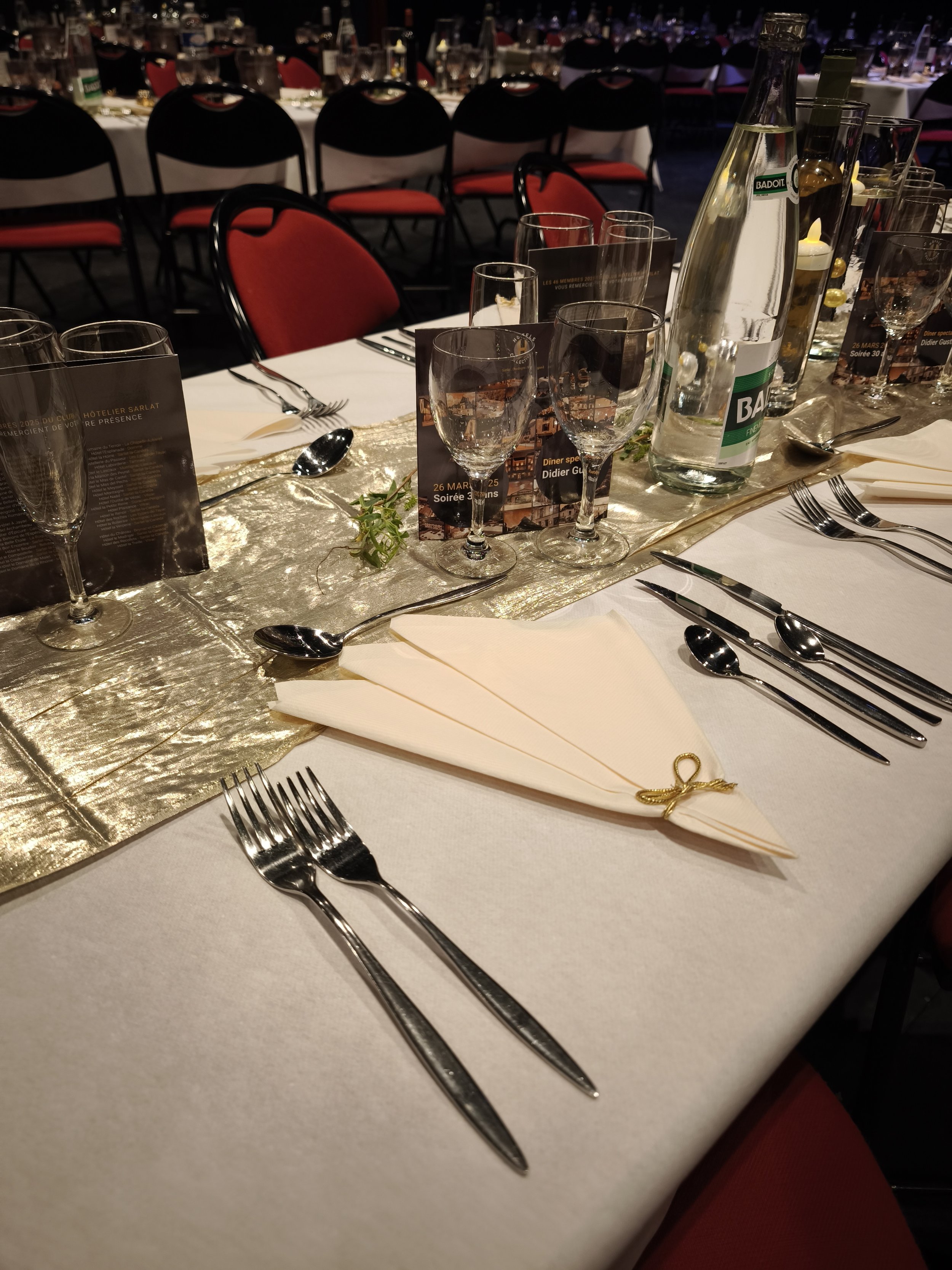 Table de banquet élégante avec des verres, des couverts en argent, une bouteille d'eau, des menus, et une nappe dorée. Chaises avec dossier noir et assise rouge en arrière-plan.