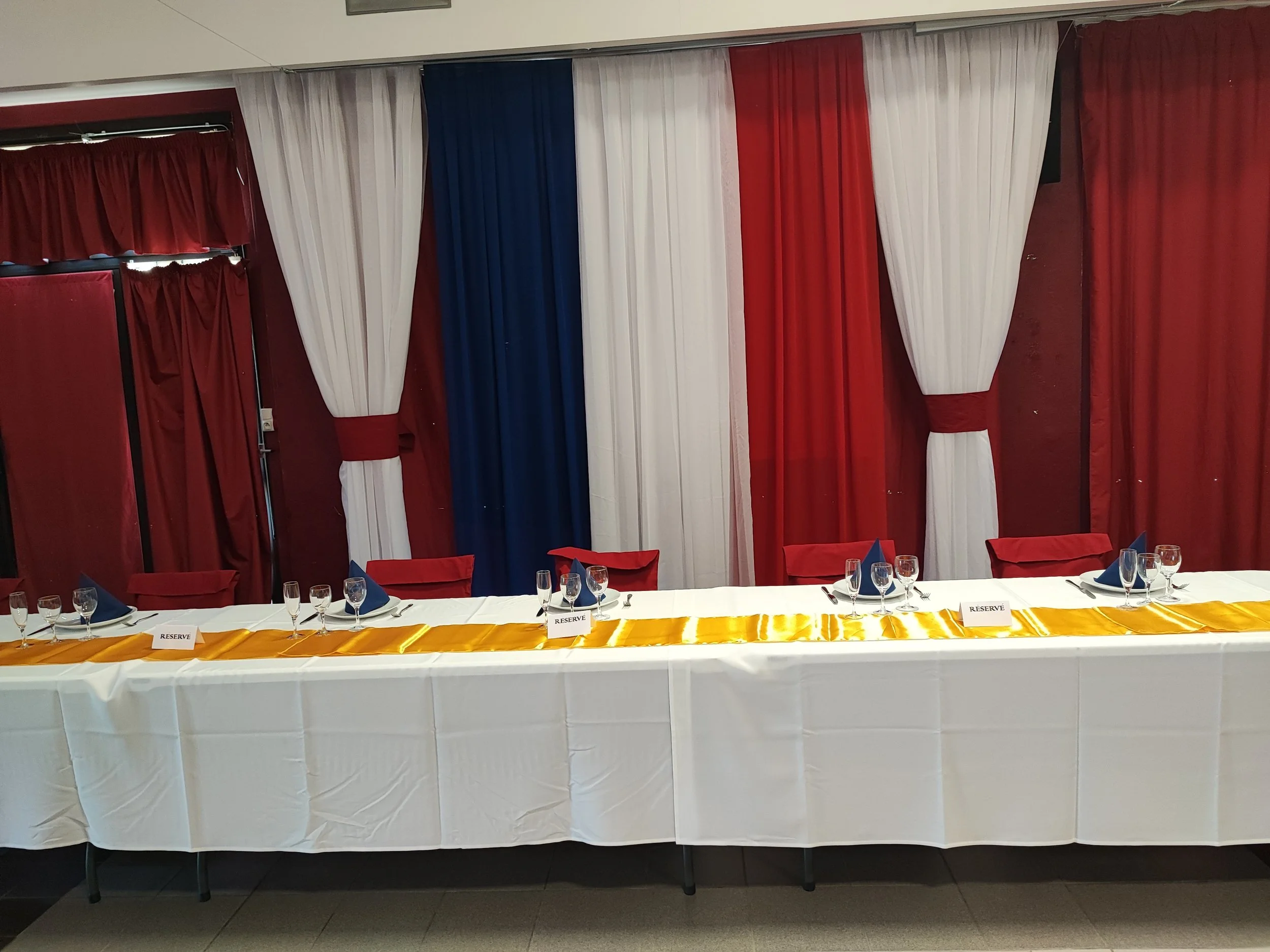 Une table décorée pour un événement avec des couverts, des verres et des badges 'Réservé' en français. En arrière-plan, des rideaux rouges, blancs et bleus ornent un mur, suggérant un décor festif ou officiel.