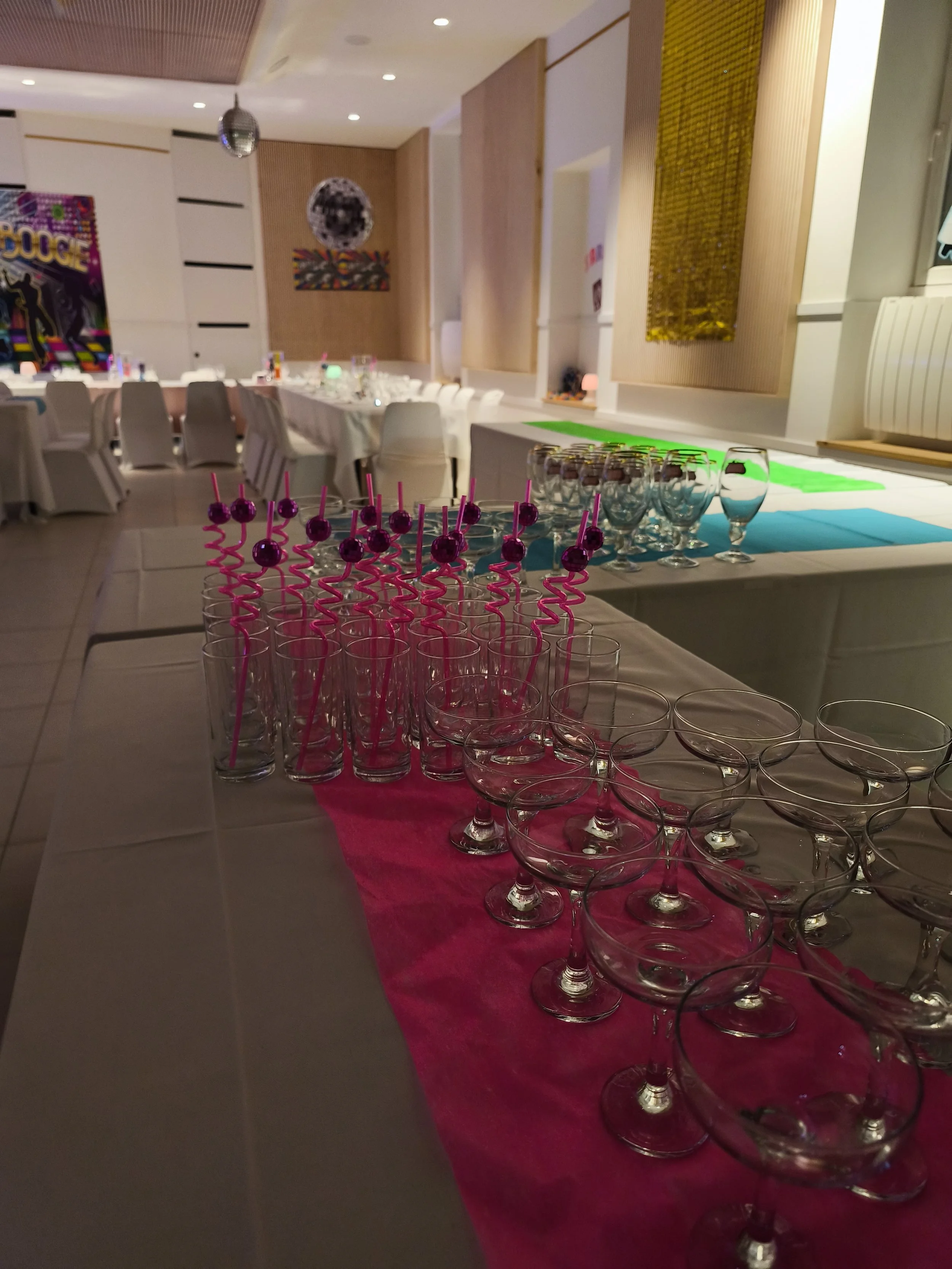 Une salle décorée pour une fête ou un événement, avec des tables, des verres, des décorations colorées, y compris des pailles roses, et un fond avec des œuvres d'art murales.