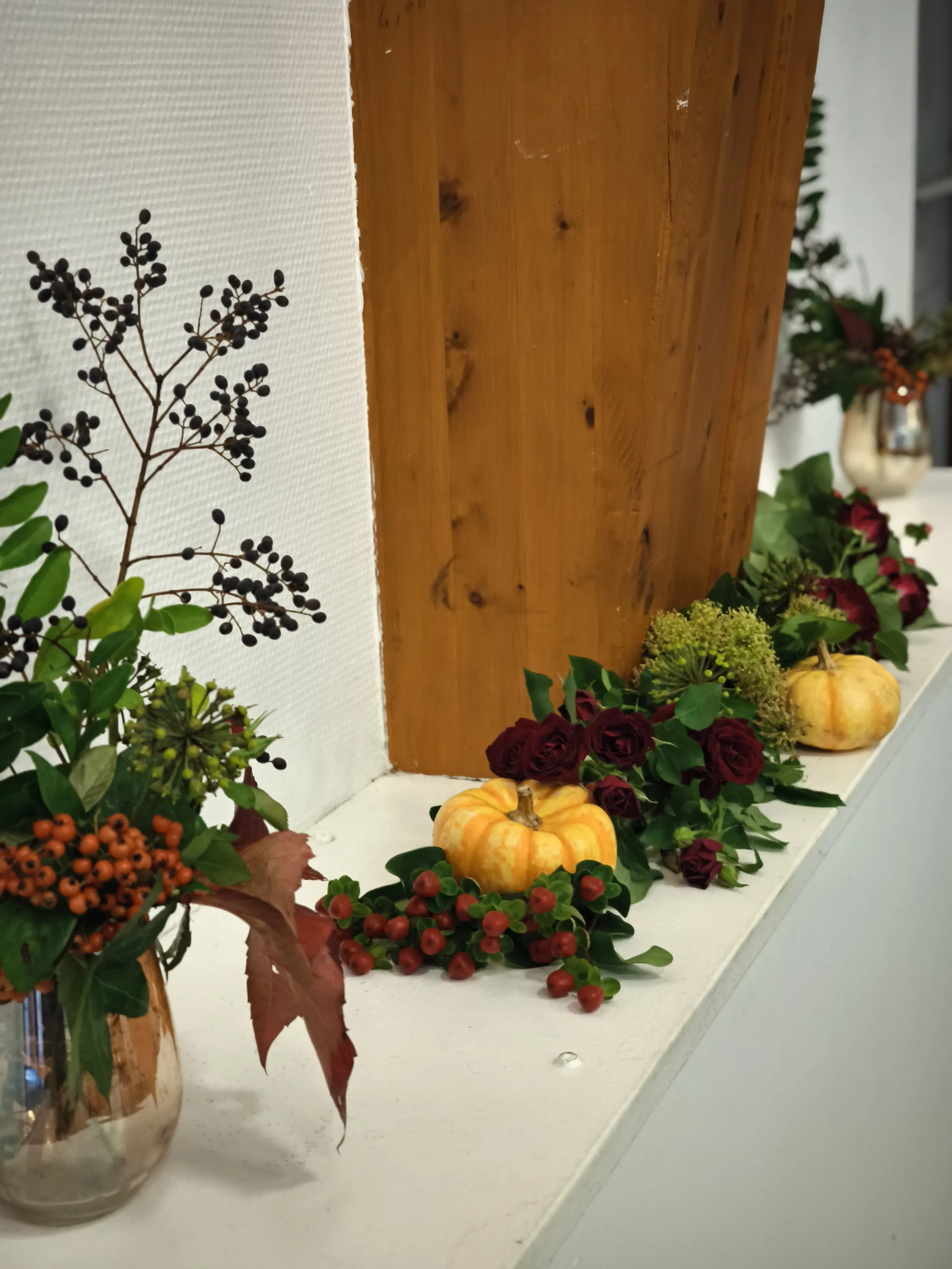 Décoration florale automnale avec de petites citrouilles, des fleurs rouges, vertes et oranges, et des branches avec feuilles ou baies, sur un rebord blanc.