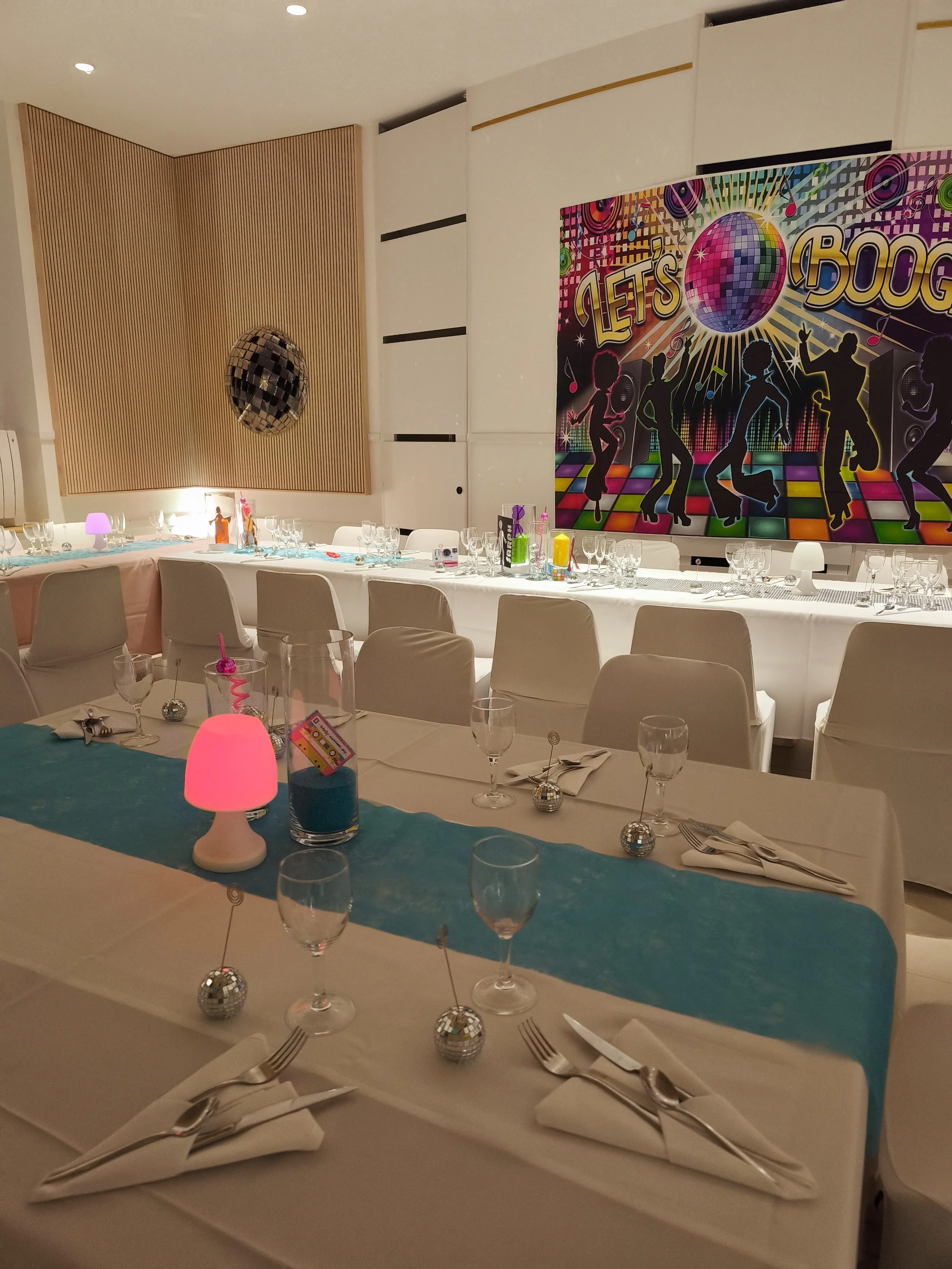 Salle de fête décorée avec une bannière colorée représentant des silhouettes de personnes dansant avec un disque en miroir et des éléments de fête. Table élégante avec nappes, verres, couverts, lampes roses et petits ballons Disco.
