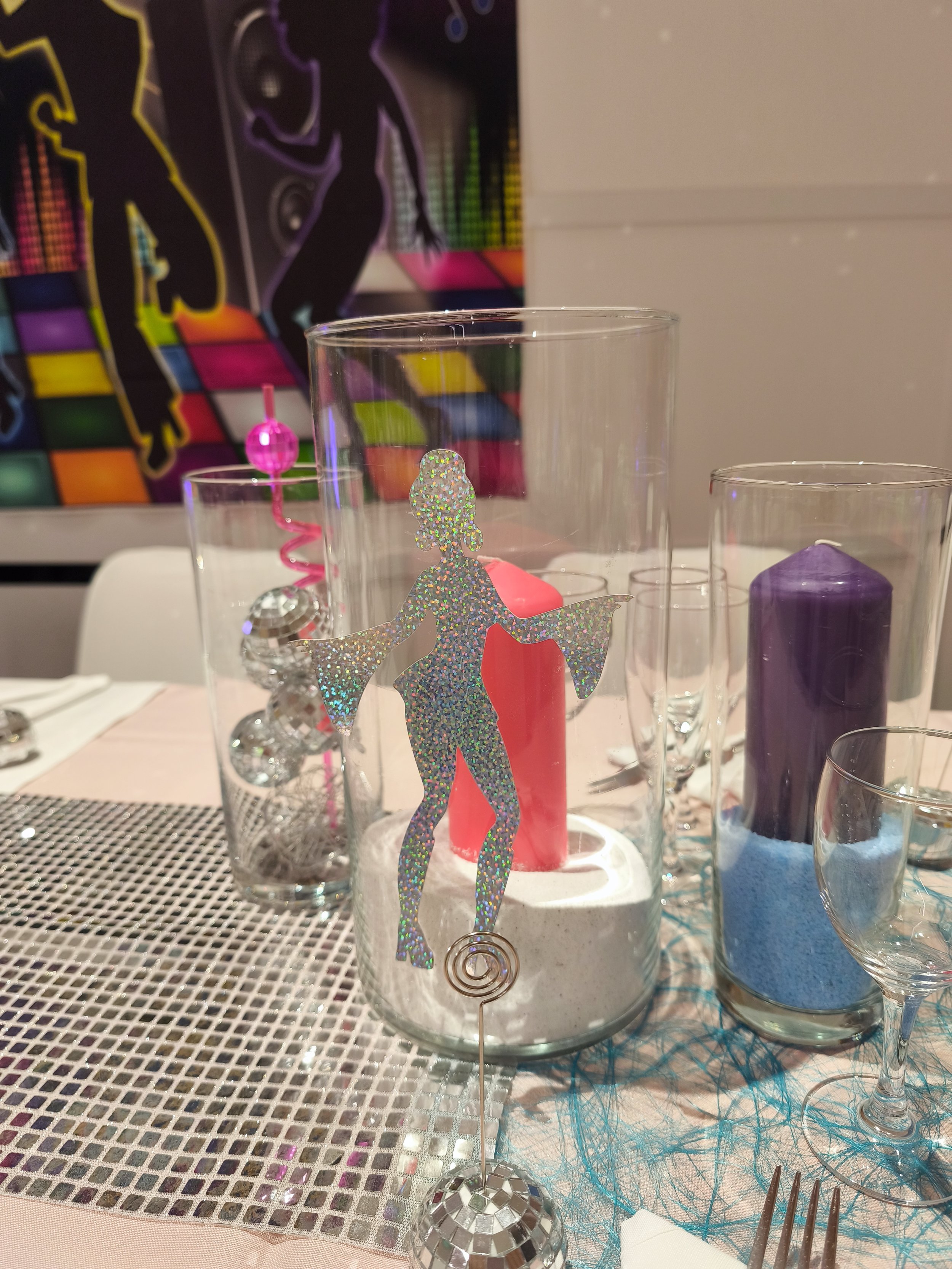 Décor de table festif avec des verres transparents, bougie violette, figurine en papier holographique d'une femme avec un foulard, napperon en fil bleu, et décorations multicolores en arrière-plan